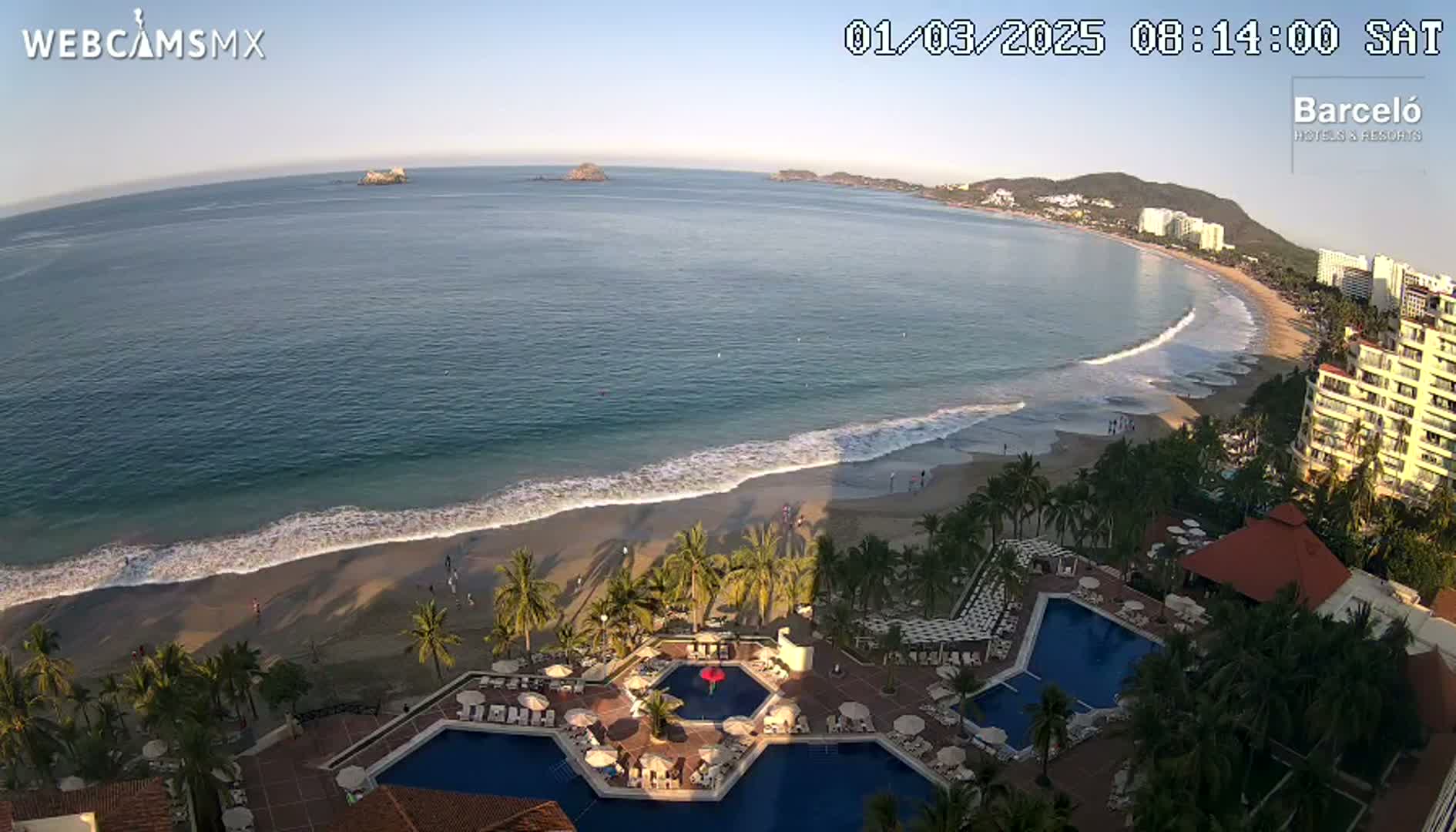 An aerial view shows a calm ocean lapping a sandy beach, with palm trees and resort pools in the foreground under a clear sky.