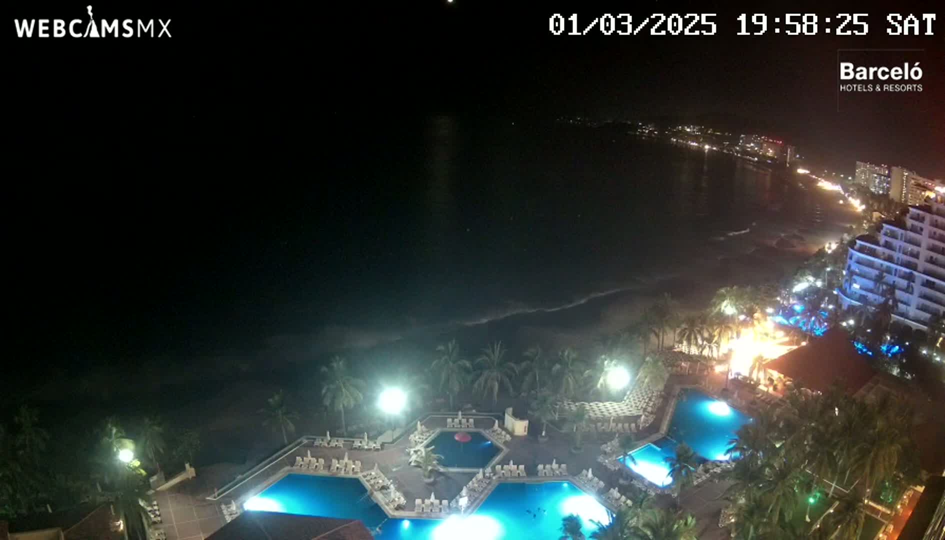 An aerial nighttime view of a resort with multiple illuminated pools, palm trees, and a dark ocean with waves visible in the distance.