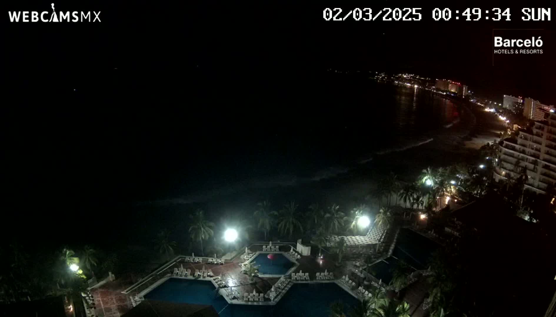 A nighttime view of a resort complex with multiple pools, illuminated by artificial lights, overlooking a dark ocean and a dimly lit coastline.