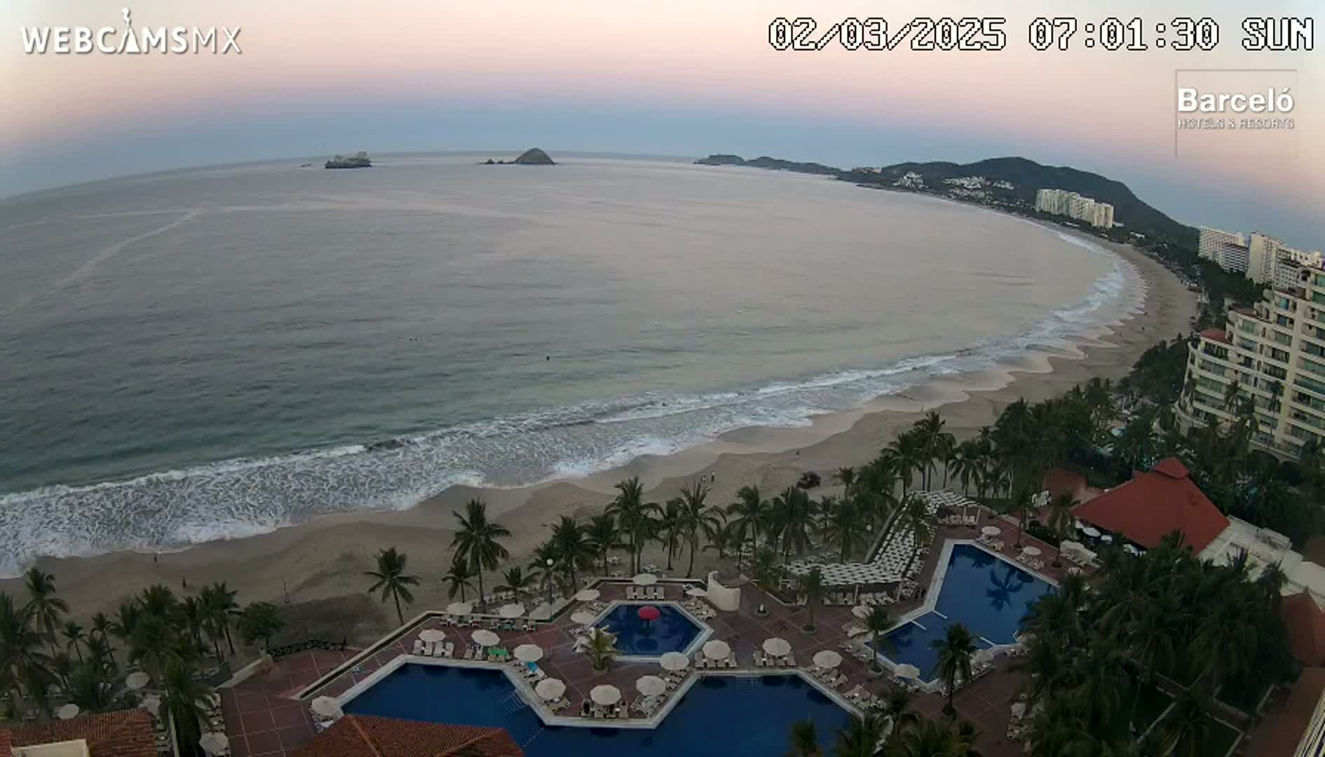 An aerial view shows a resort with multiple pools next to a sandy beach and calm ocean under a twilight sky.
