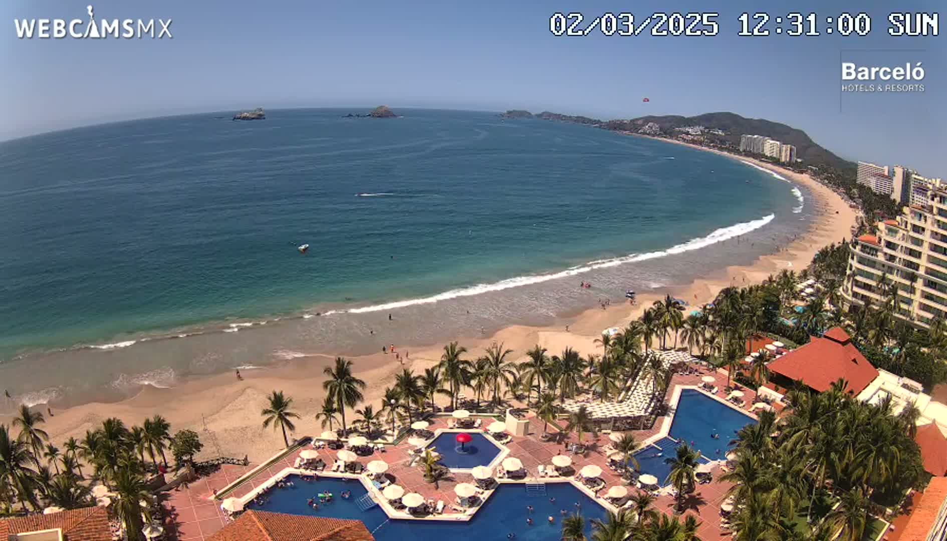 An aerial view shows a resort with multiple pools next to a sandy beach and calm, blue ocean under a clear, sunny sky.
