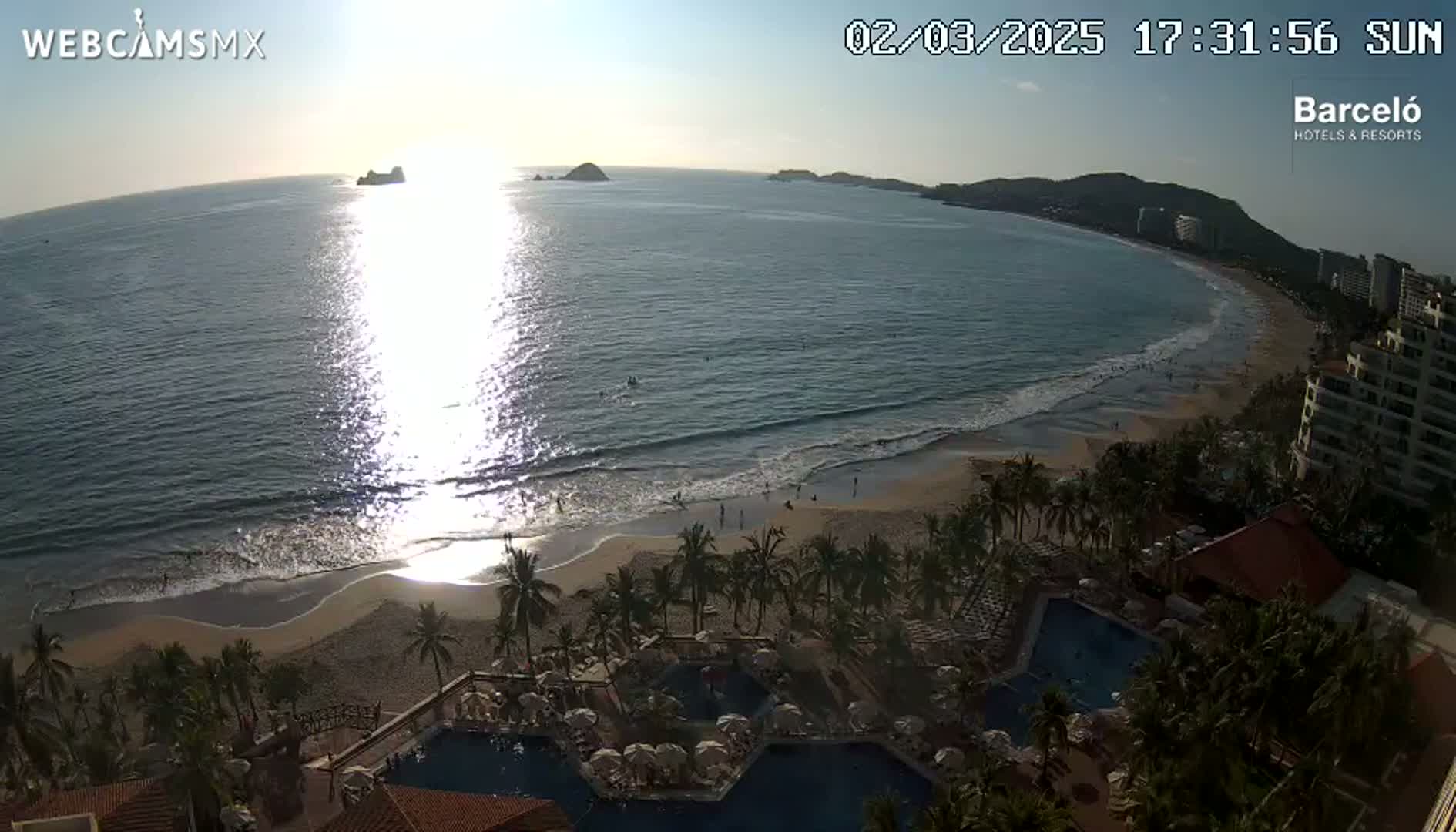 A beach with palm trees and resort pools is bathed in the late afternoon sun, with calm waves washing ashore a sandy beach, and islands visible in the distance.