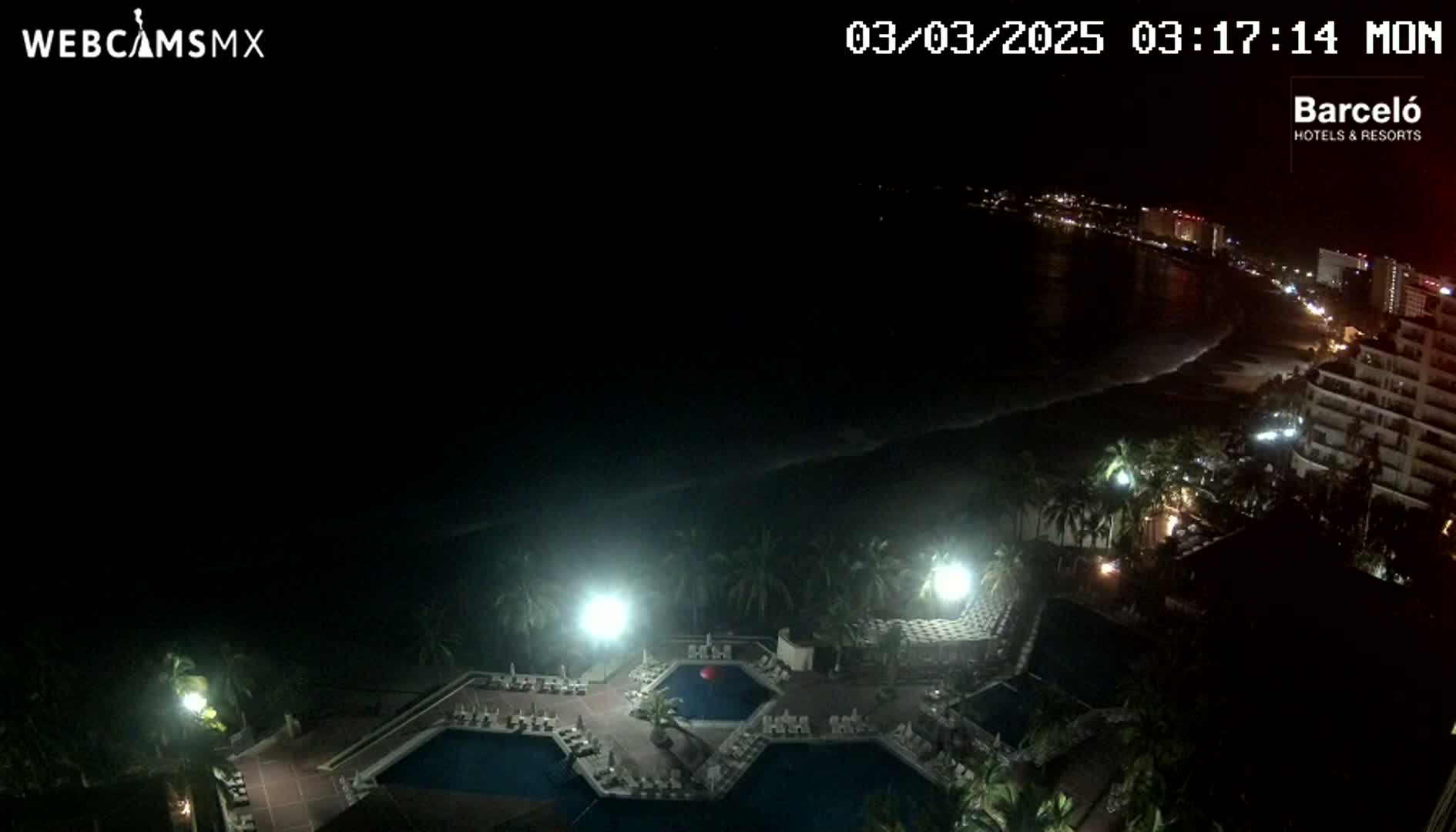 A nighttime aerial view shows a resort complex with illuminated pools and surrounding buildings, overlooking a dark ocean with a faintly visible shoreline and city lights in the distance.