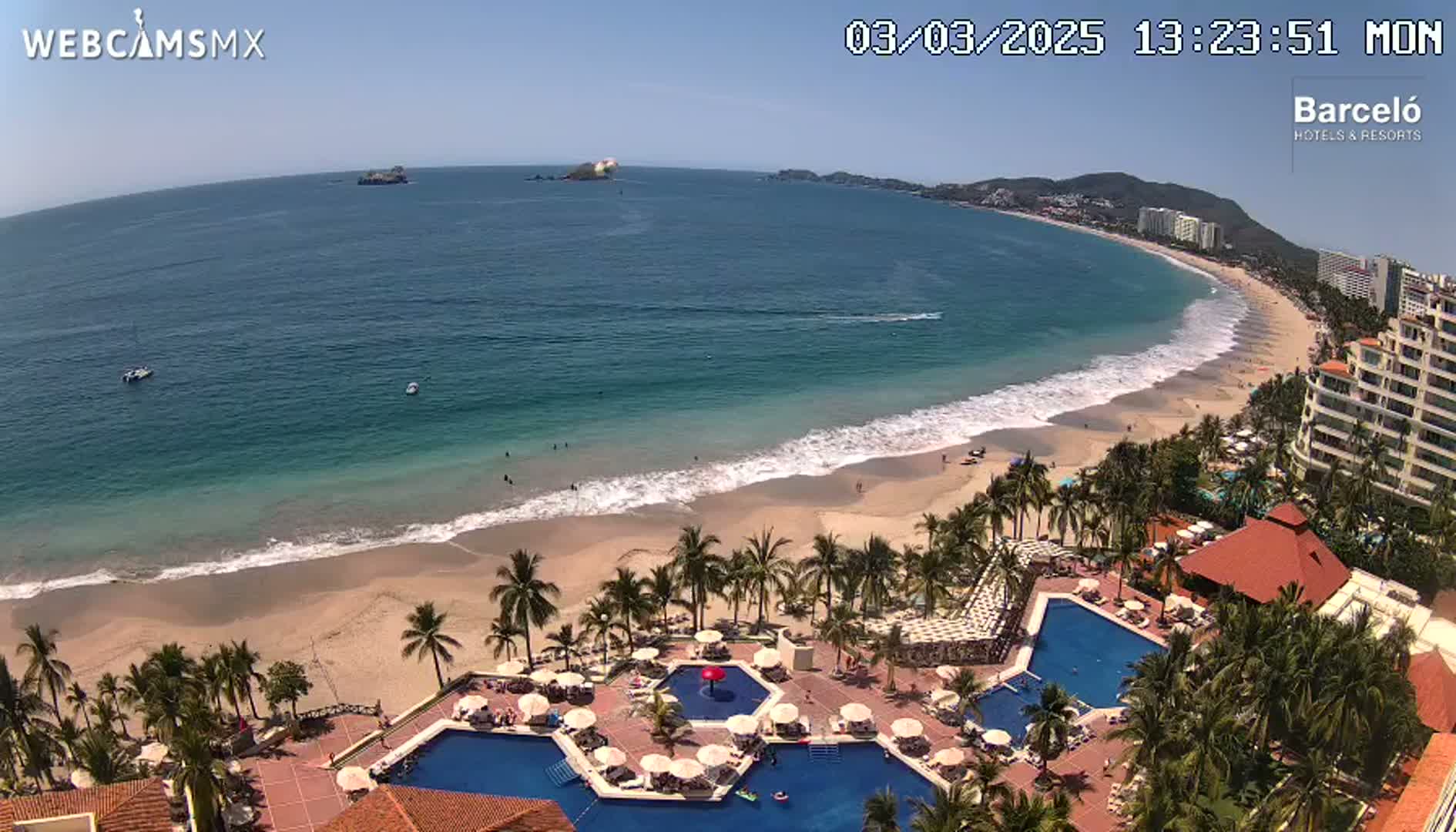 An aerial view shows a beach with gentle waves under a clear blue sky, featuring a resort with multiple pools and palm trees.
