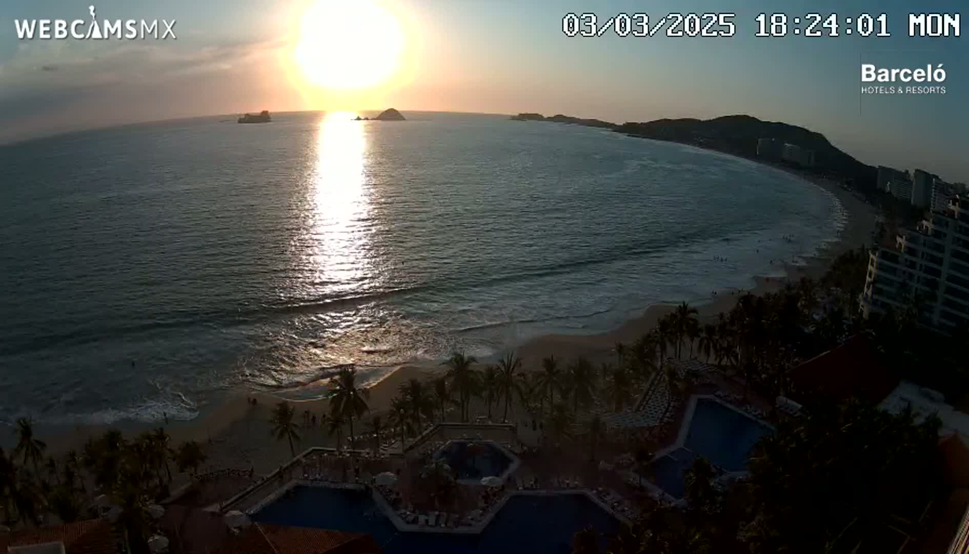 An ocean sunset casts a shimmering path across the water towards a sandy beach lined with palm trees and a resort with multiple pools.