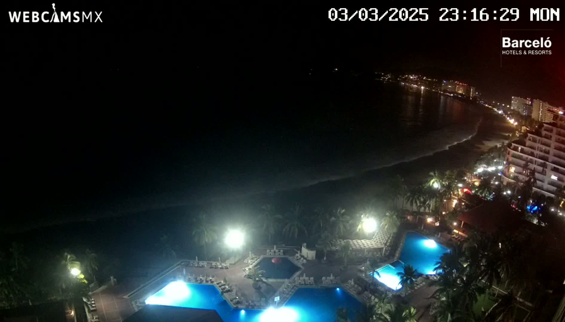 At night, a resort with illuminated pools is situated near a dark ocean beach and a city skyline under a clear sky.
