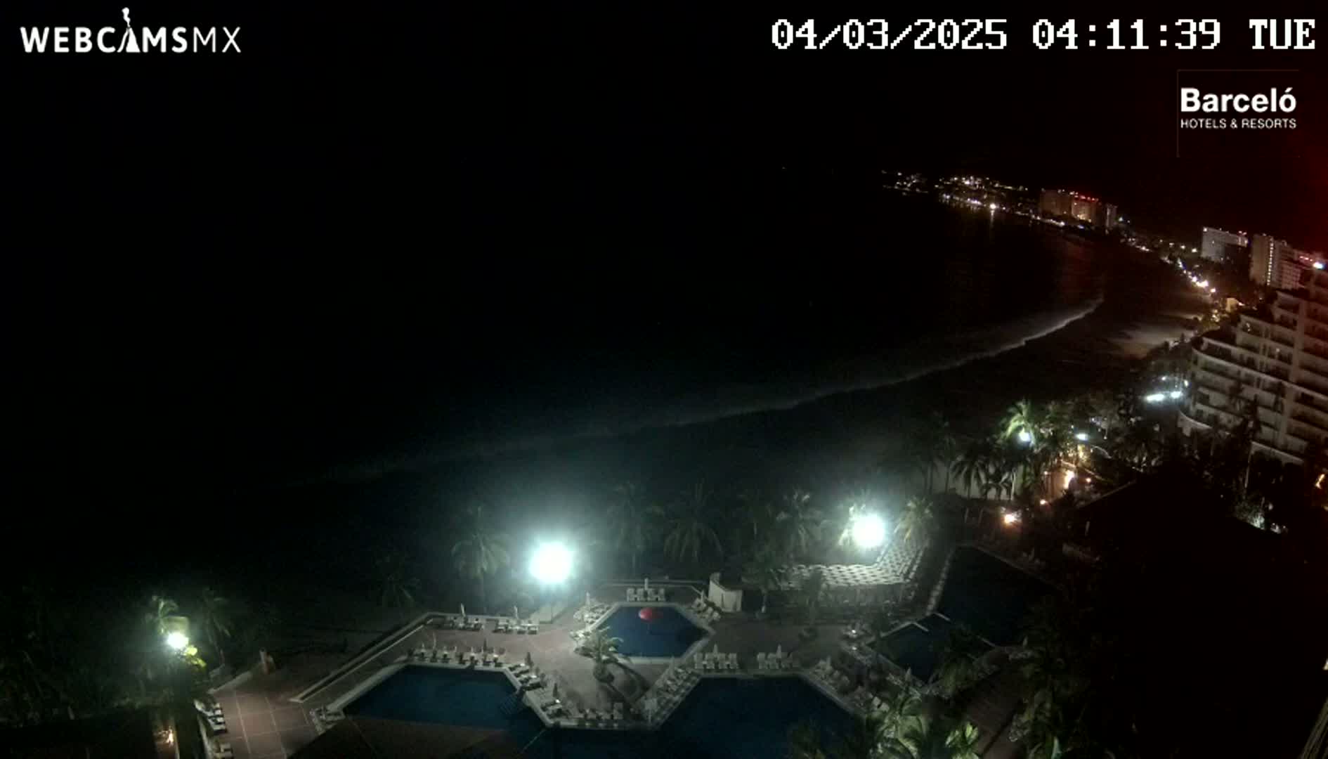 A nighttime view of a resort complex with multiple pools, illuminated by lights, overlooking a dark ocean with a gently lapping shoreline.