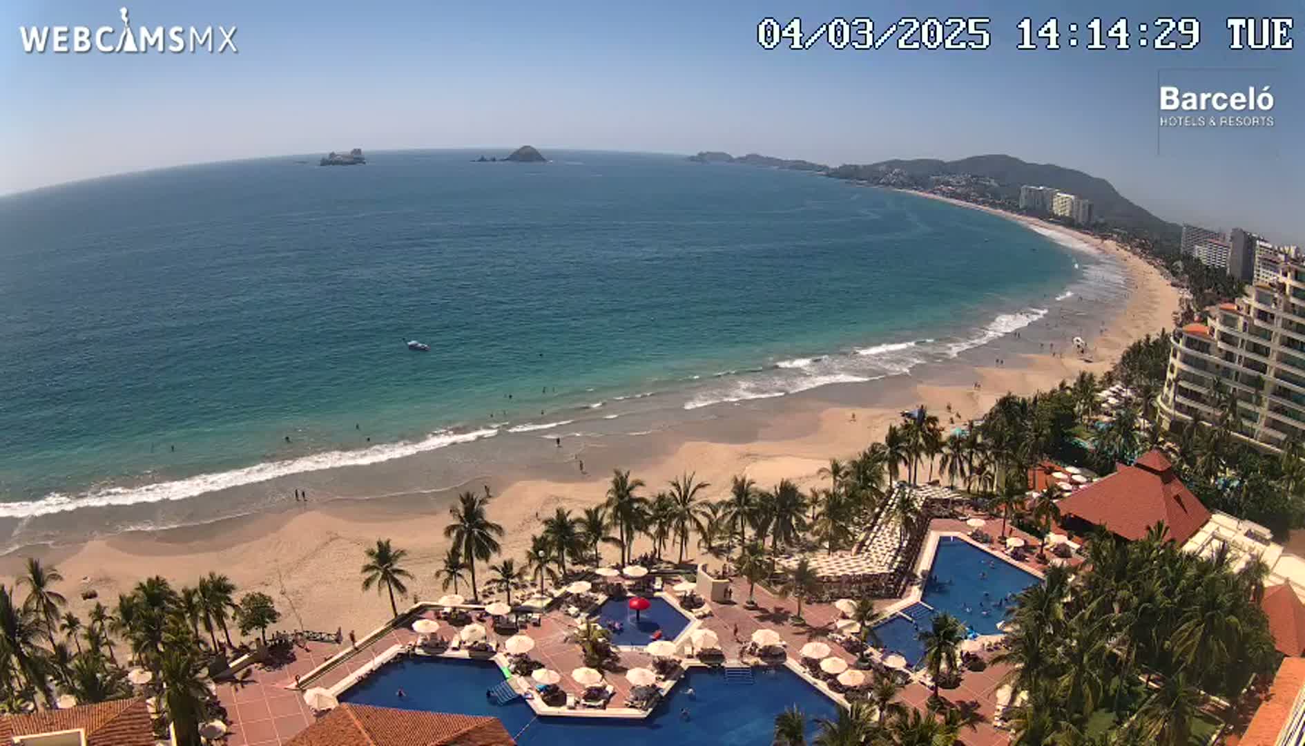 An aerial view shows a resort with multiple pools nestled among palm trees next to a sandy beach and ocean under a clear, sunny sky.