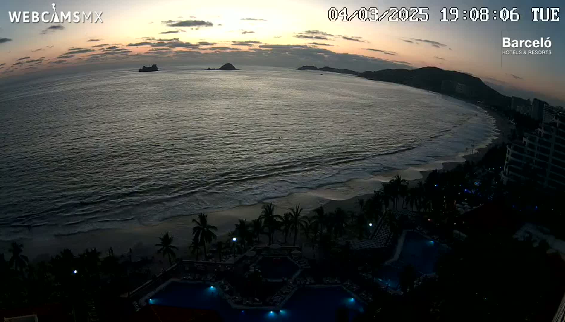 An ocean beach at sunset, with palm trees and resort pools visible along the shoreline, under a mostly clear sky.