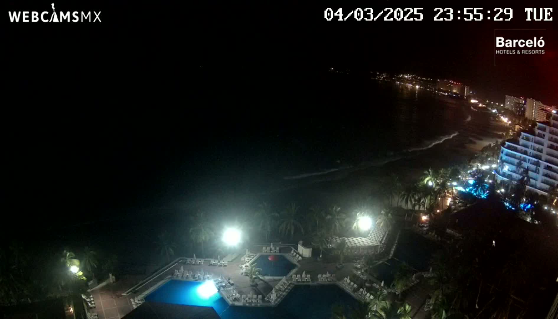 A nighttime view of a resort pool area overlooking a dark ocean with a shoreline of lights in the distance.