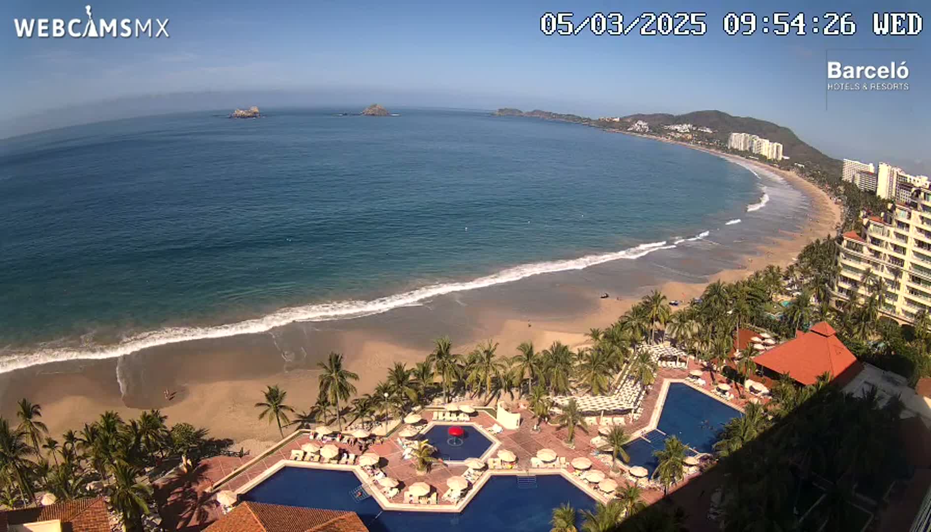 An aerial view shows a beach with calm ocean waves under a clear blue sky, featuring a resort with multiple pools and palm trees lining the shore.