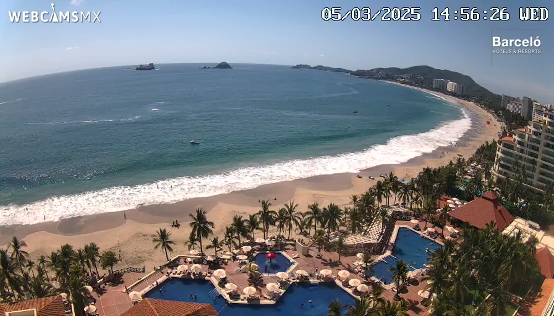 An aerial view shows a resort with multiple pools overlooking a sandy beach with waves lapping the shore under a sunny sky.