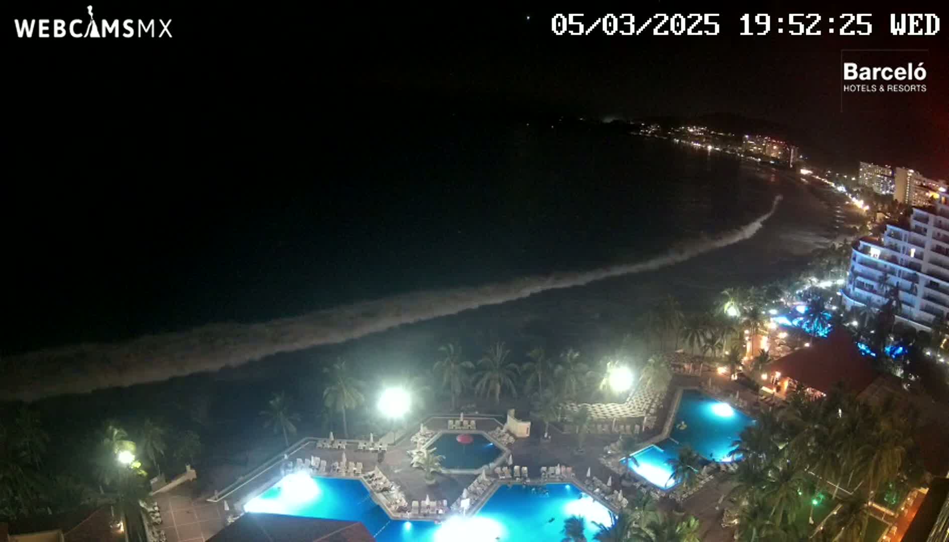 A nighttime view of a resort with multiple illuminated pools, palm trees, and a coastline with gently crashing waves under a dark sky.