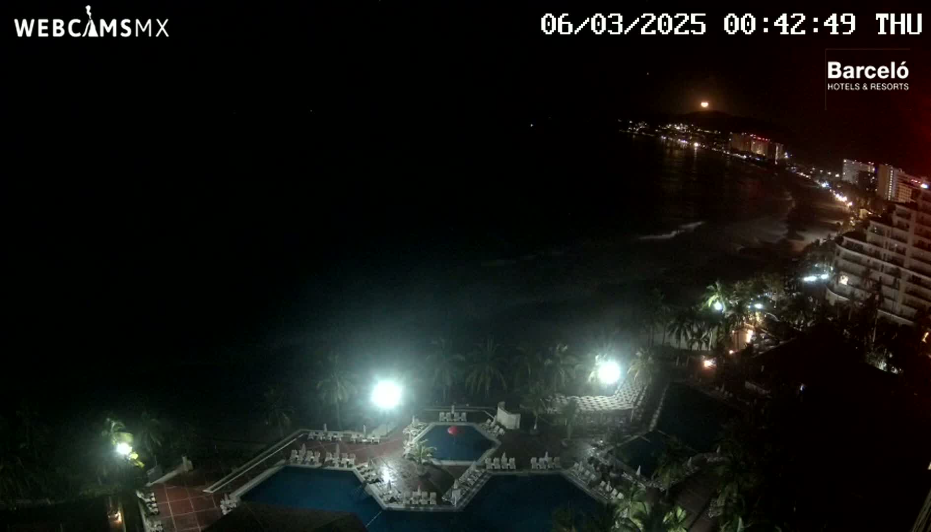 A nighttime, overhead view of a resort pool area with a brightly lit beach and ocean visible in the distance under a clear sky with a visible moon.