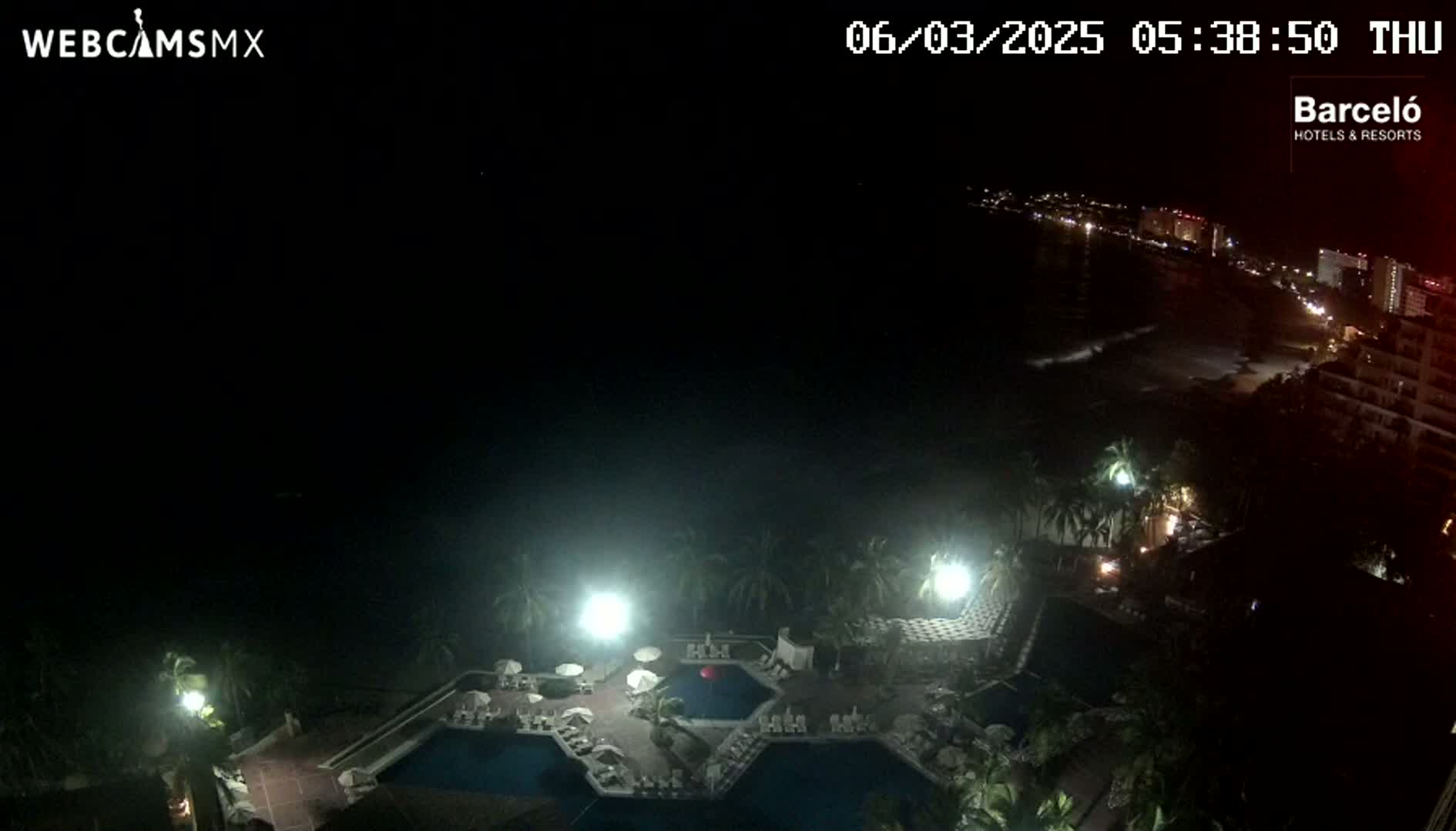 A nighttime aerial view shows a resort pool area with several pools illuminated, and a dark ocean with a city skyline visible in the distance under a clear, dark sky.
