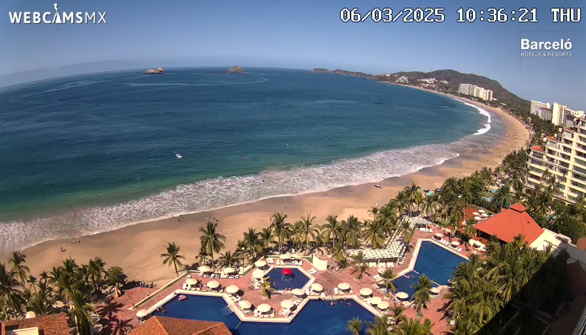 A beach with calm waves and a resort with multiple pools is seen from an elevated viewpoint on a sunny day.