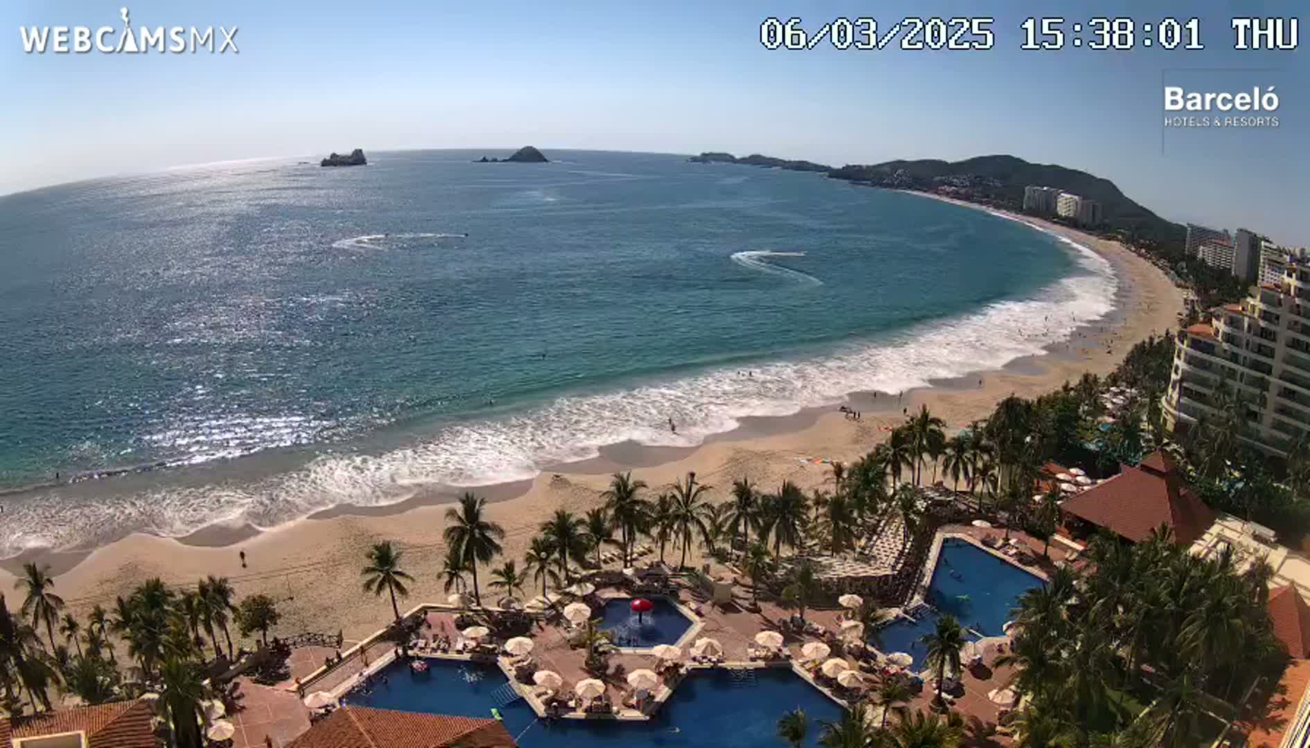 A beach with gentle waves under a clear blue sky is seen from above, showcasing a resort with multiple pools surrounded by palm trees.