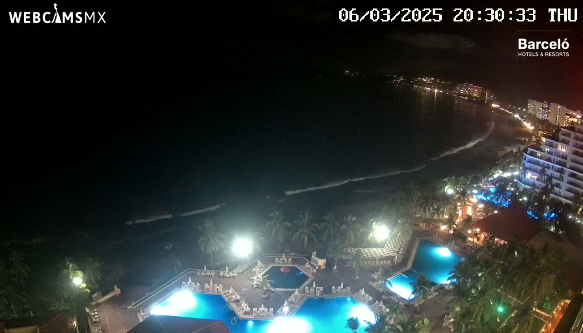 A nighttime, high-angle view shows a resort with illuminated pools overlooking a dark ocean with gentle waves under a clear, starless sky.