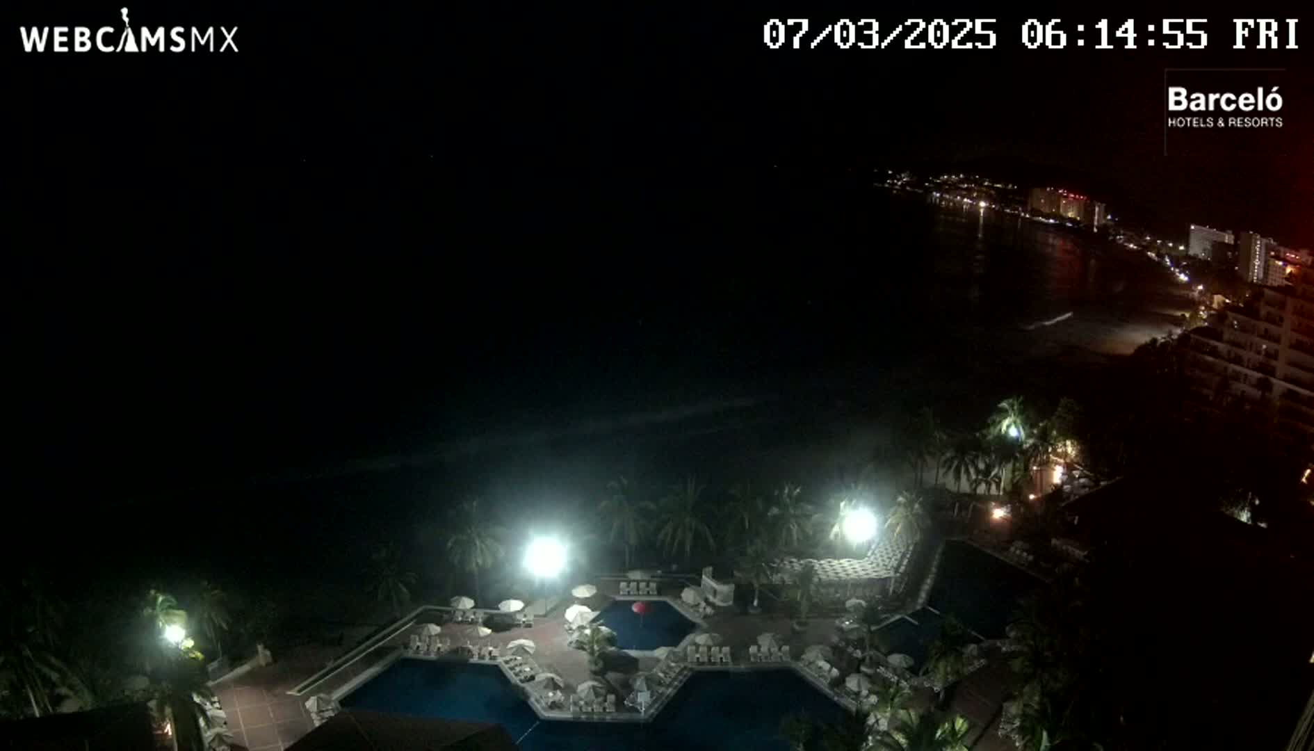 A nighttime aerial view of a resort pool area with palm trees, illuminated by artificial lights, overlooking a dark ocean and a city skyline.