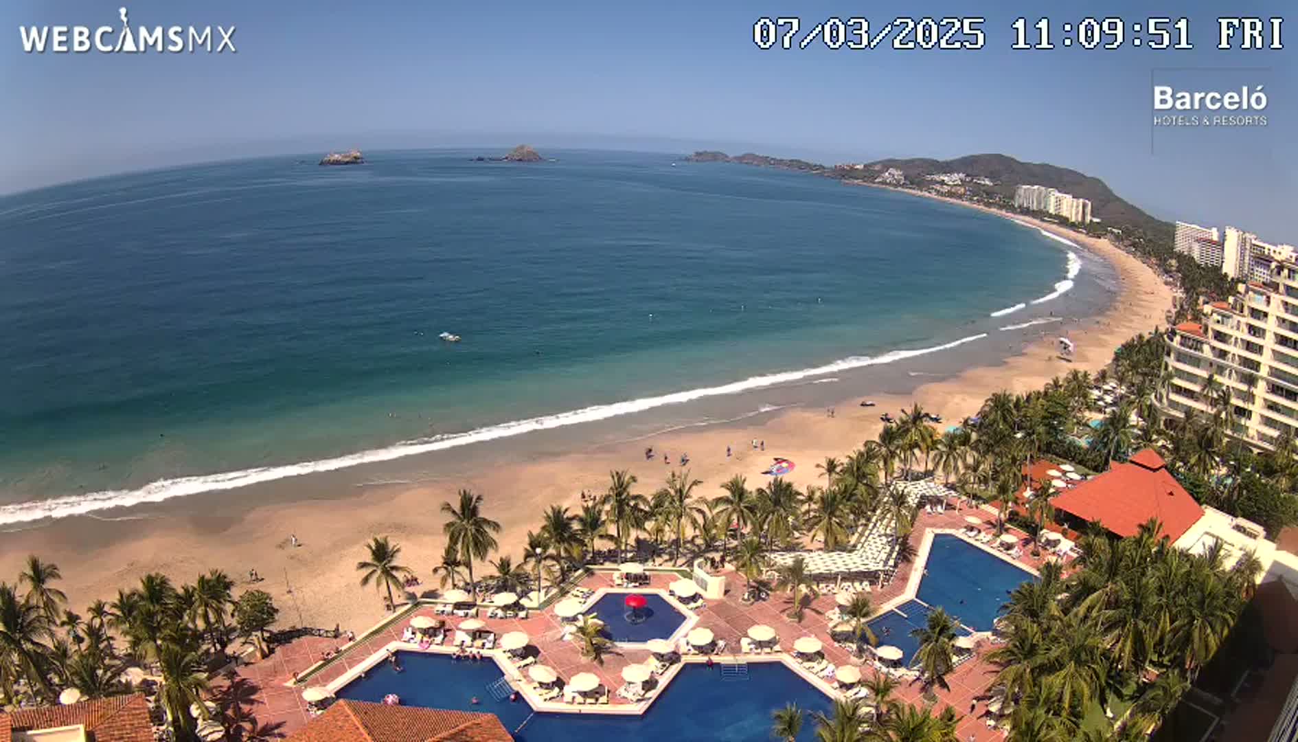 A beach resort with multiple pools is situated on a sandy beach next to a calm, blue ocean under a clear, sunny sky.