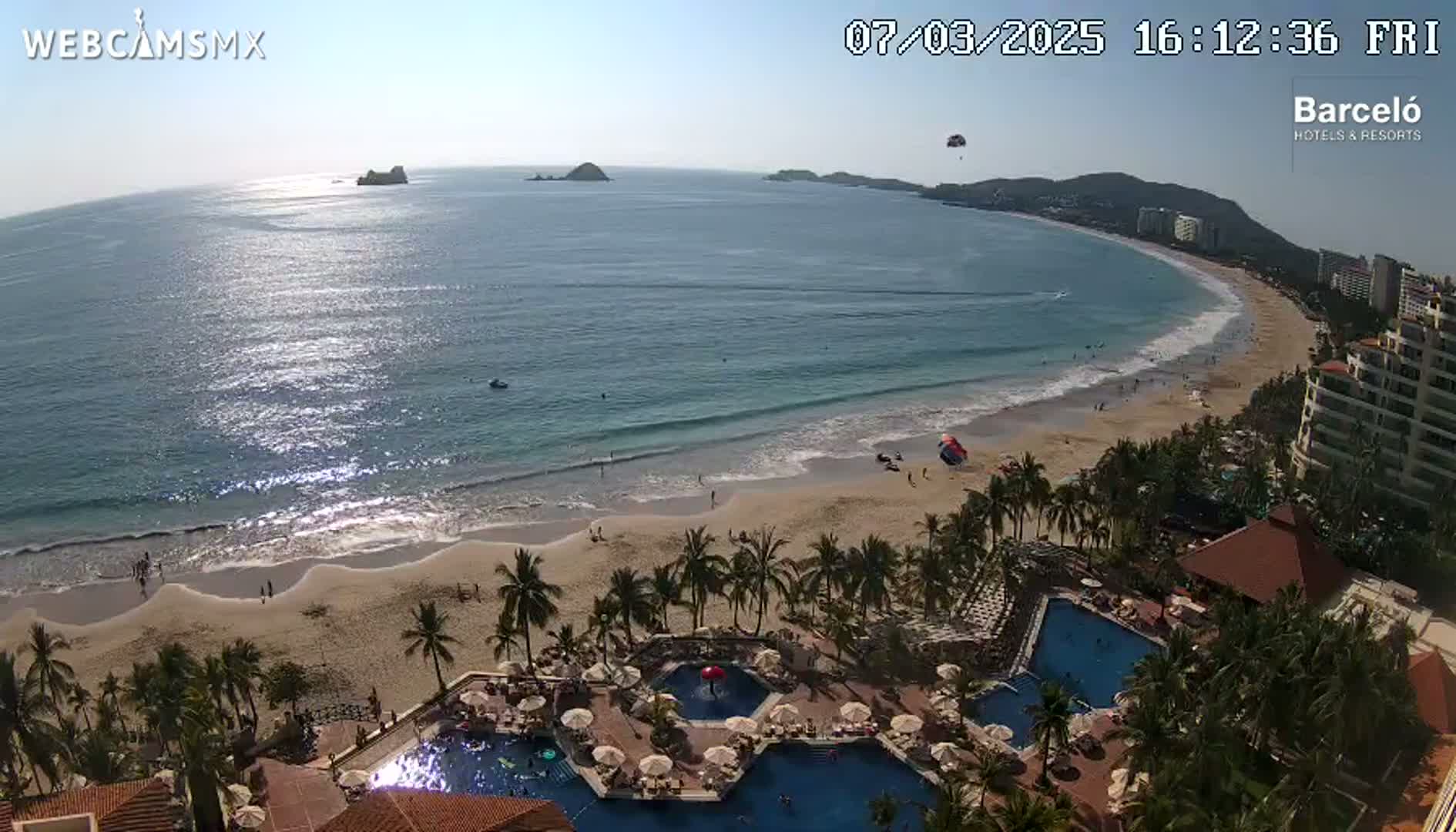 An aerial view shows a beach with calm, clear water under a sunny sky, adjacent to a resort with multiple pools and palm trees.