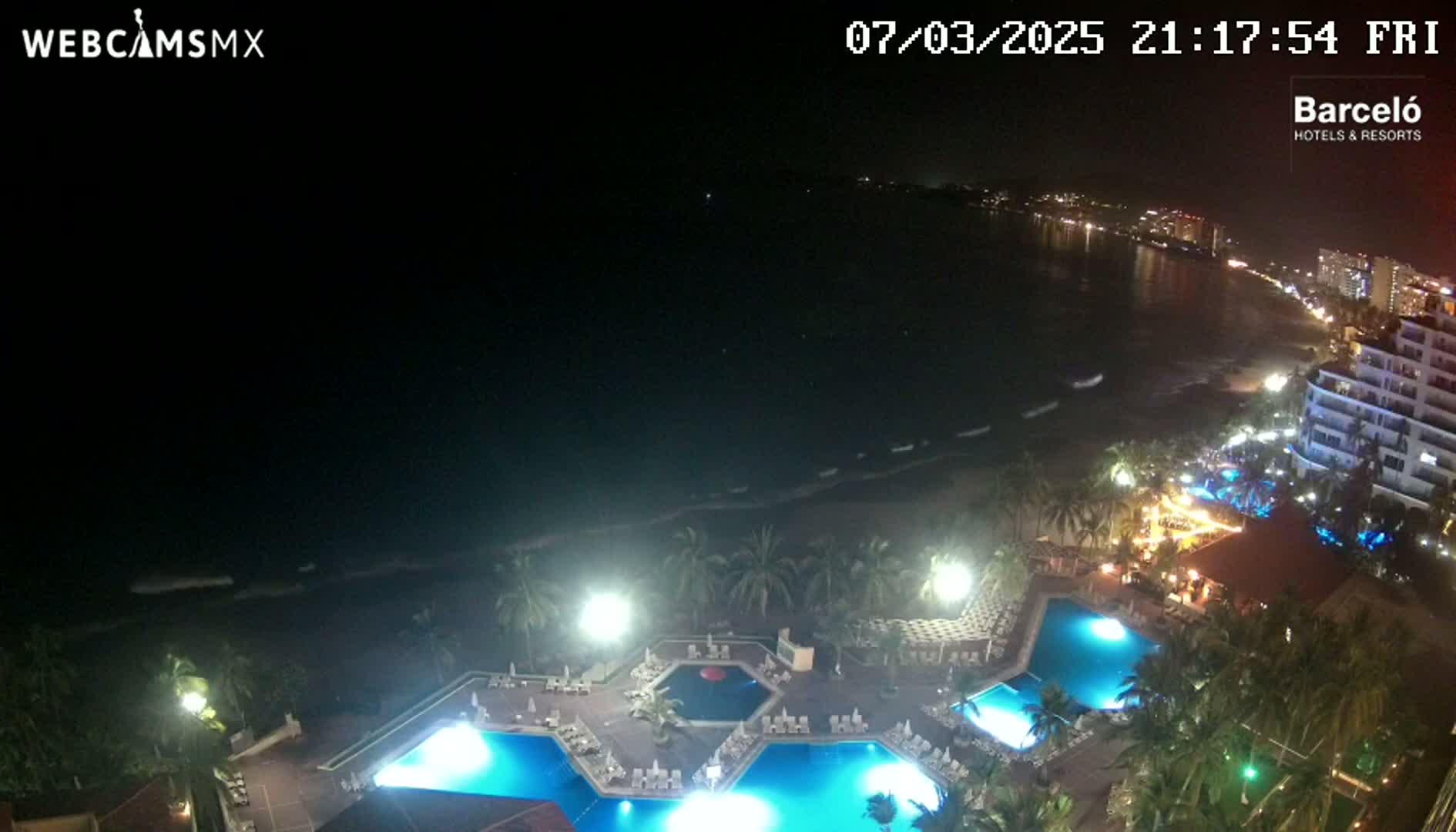 An aerial nighttime view of a resort with illuminated pools overlooking a dark ocean with gentle waves under a clear sky.