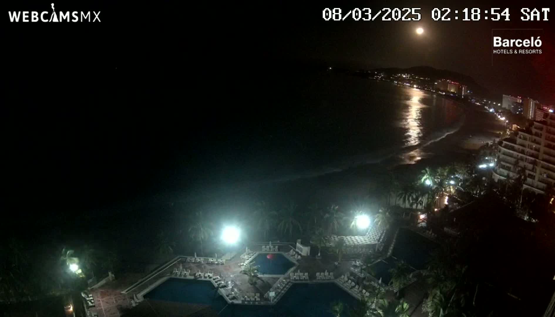 A nighttime view of a resort with multiple pools, illuminated by artificial lights, overlooks a moonlit ocean beach with a city skyline visible in the distance.