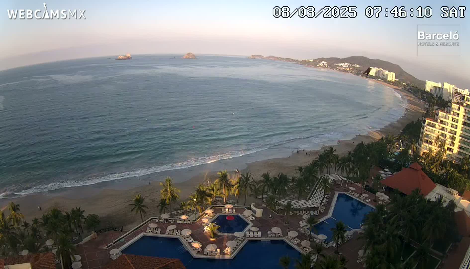 An aerial view shows a calm ocean with a sandy beach, palm trees, and a resort with multiple pools at sunrise.