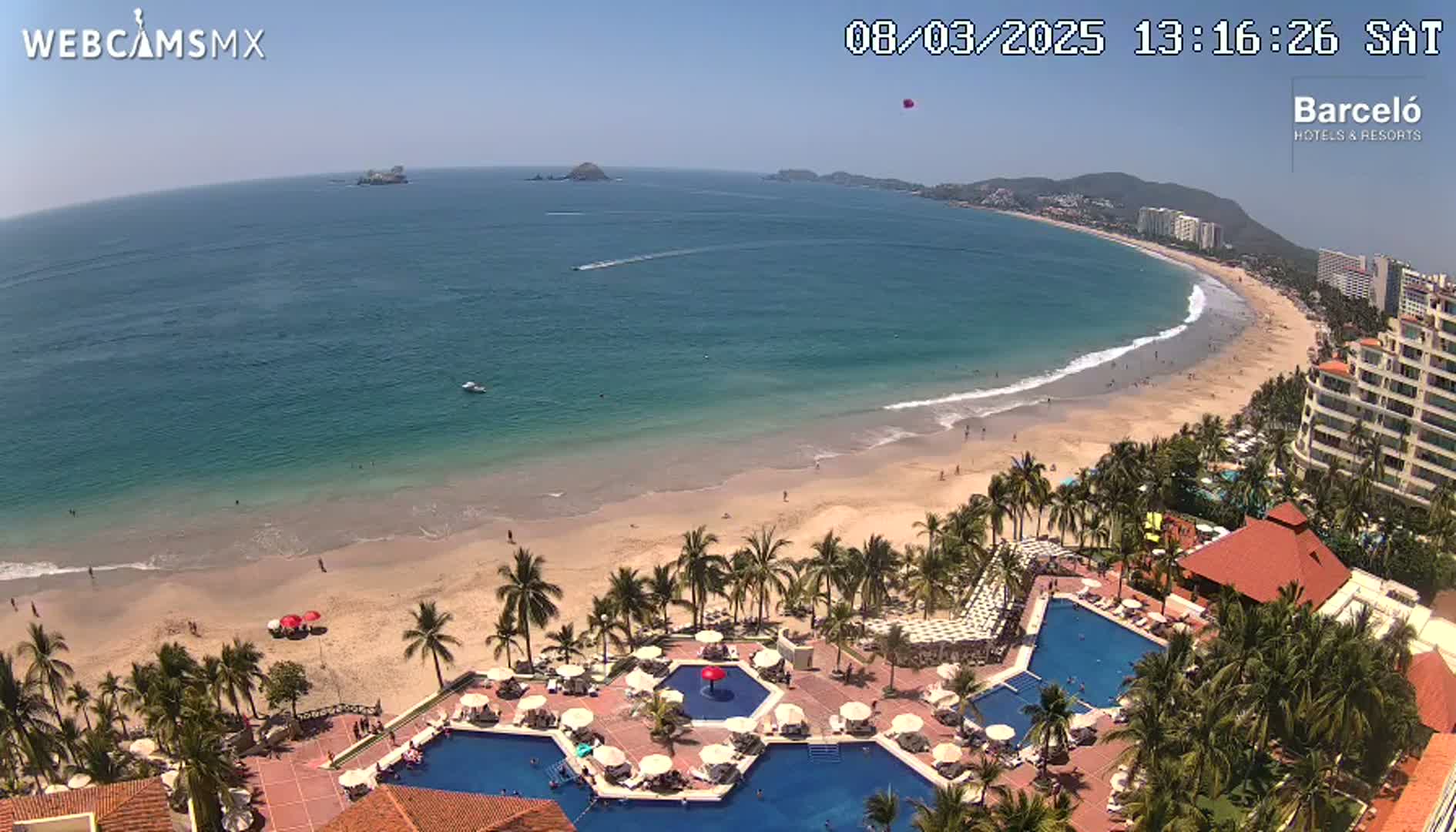An aerial view shows a resort with multiple pools overlooking a sandy beach and calm ocean on a sunny day.
