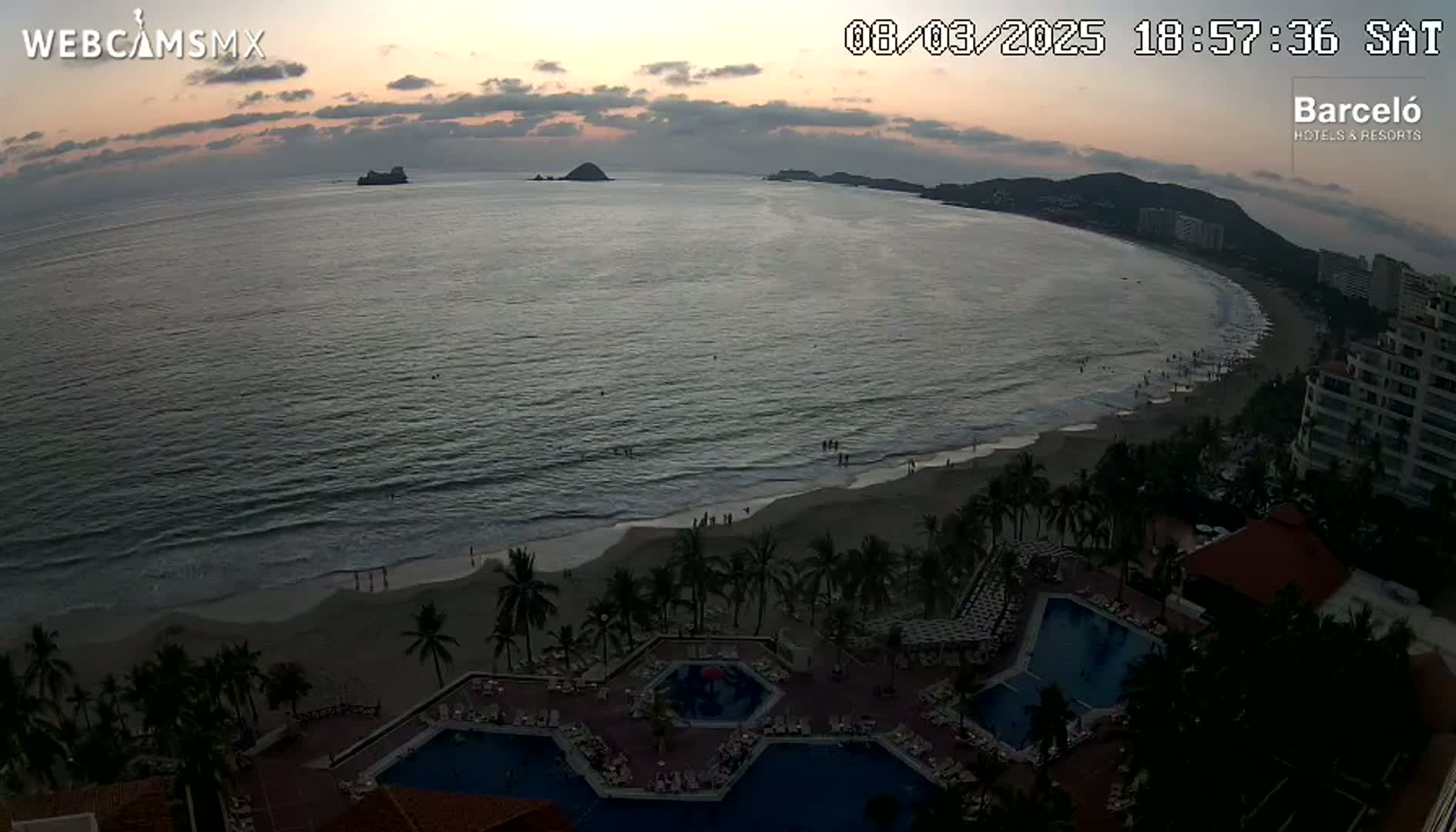 A twilight view shows a crescent-shaped beach with people on it, palm trees, resort pools, and buildings, all overlooking a calm ocean and distant islands under a partly cloudy sky.