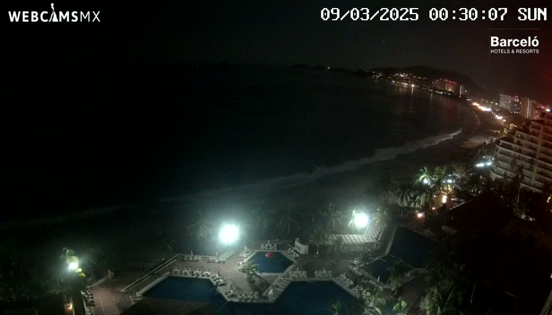 A nighttime view of a resort's pool area overlooking a dark ocean with a gently lapping shoreline and a dimly lit city in the distance.