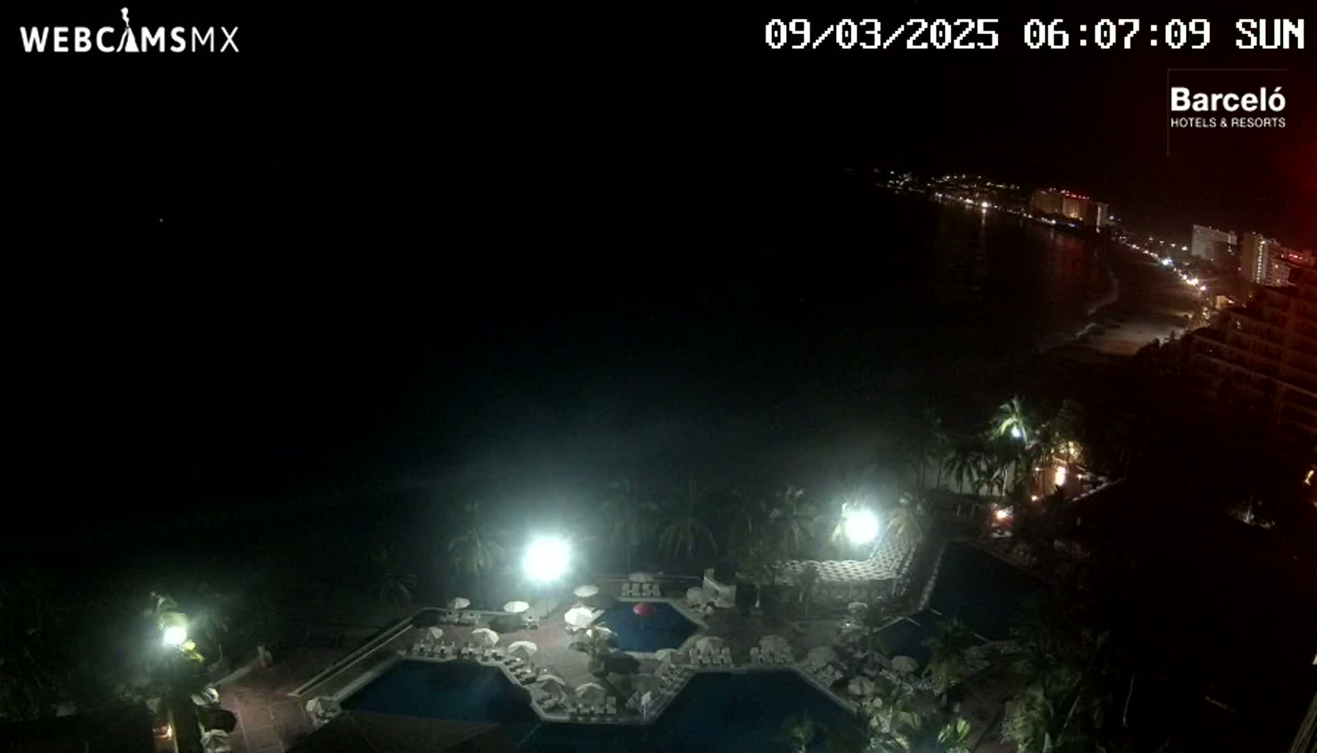 A nighttime, overhead view of a resort pool area with a dimly lit beach and coastline visible in the distance.