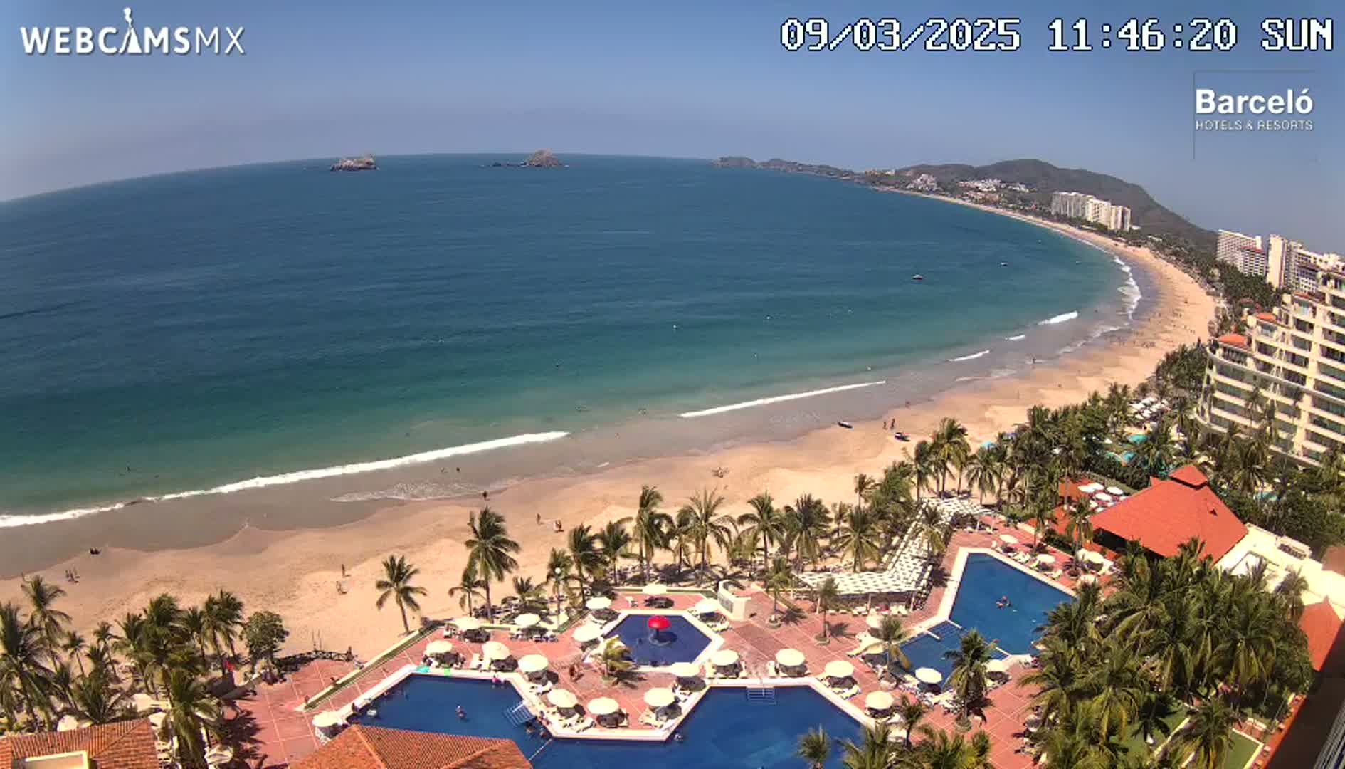 An aerial view shows a resort with multiple pools next to a sandy beach and calm ocean on a sunny day.