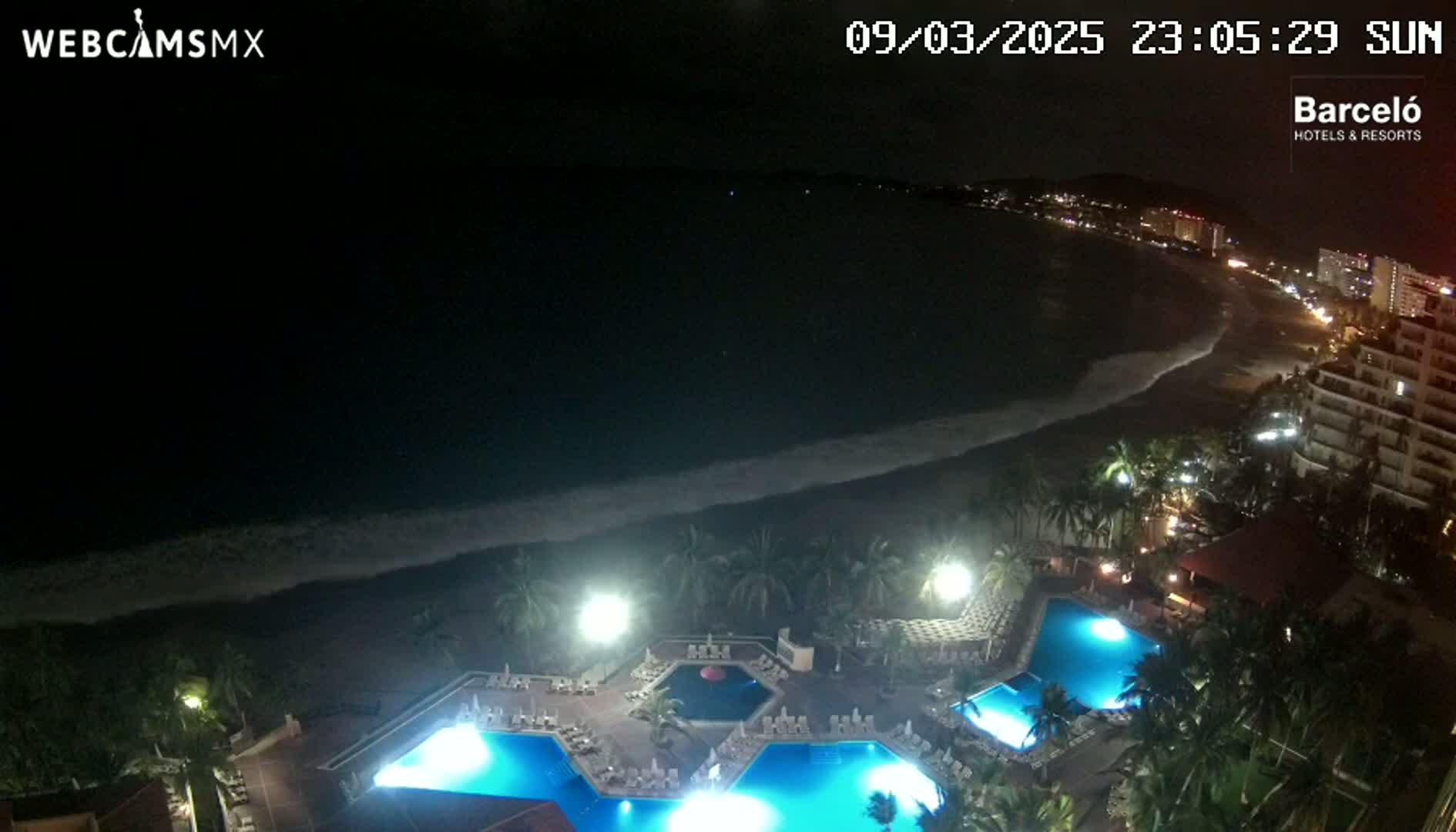 A nighttime, overhead view shows a resort with multiple illuminated pools overlooking a dark ocean with gently lapping waves and a beach lined with buildings.