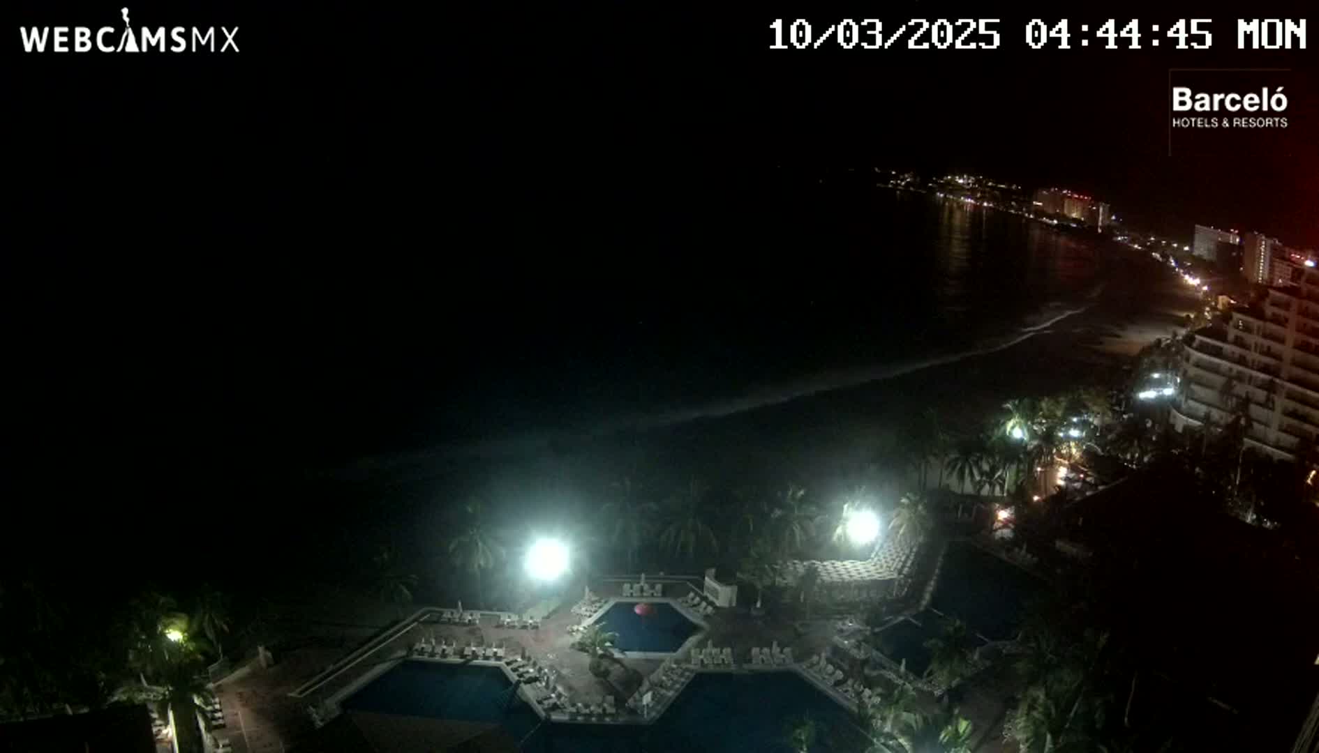 A nighttime aerial view shows a resort with illuminated pools and surrounding buildings next to a dark beach and ocean under a clear sky.
