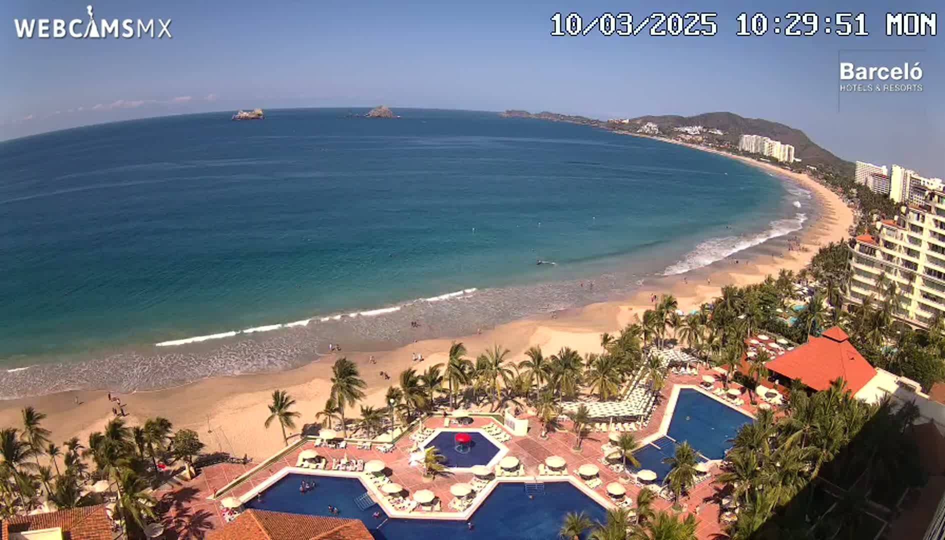 An aerial view shows a resort with multiple pools overlooking a sandy beach and calm, blue ocean on a sunny day.