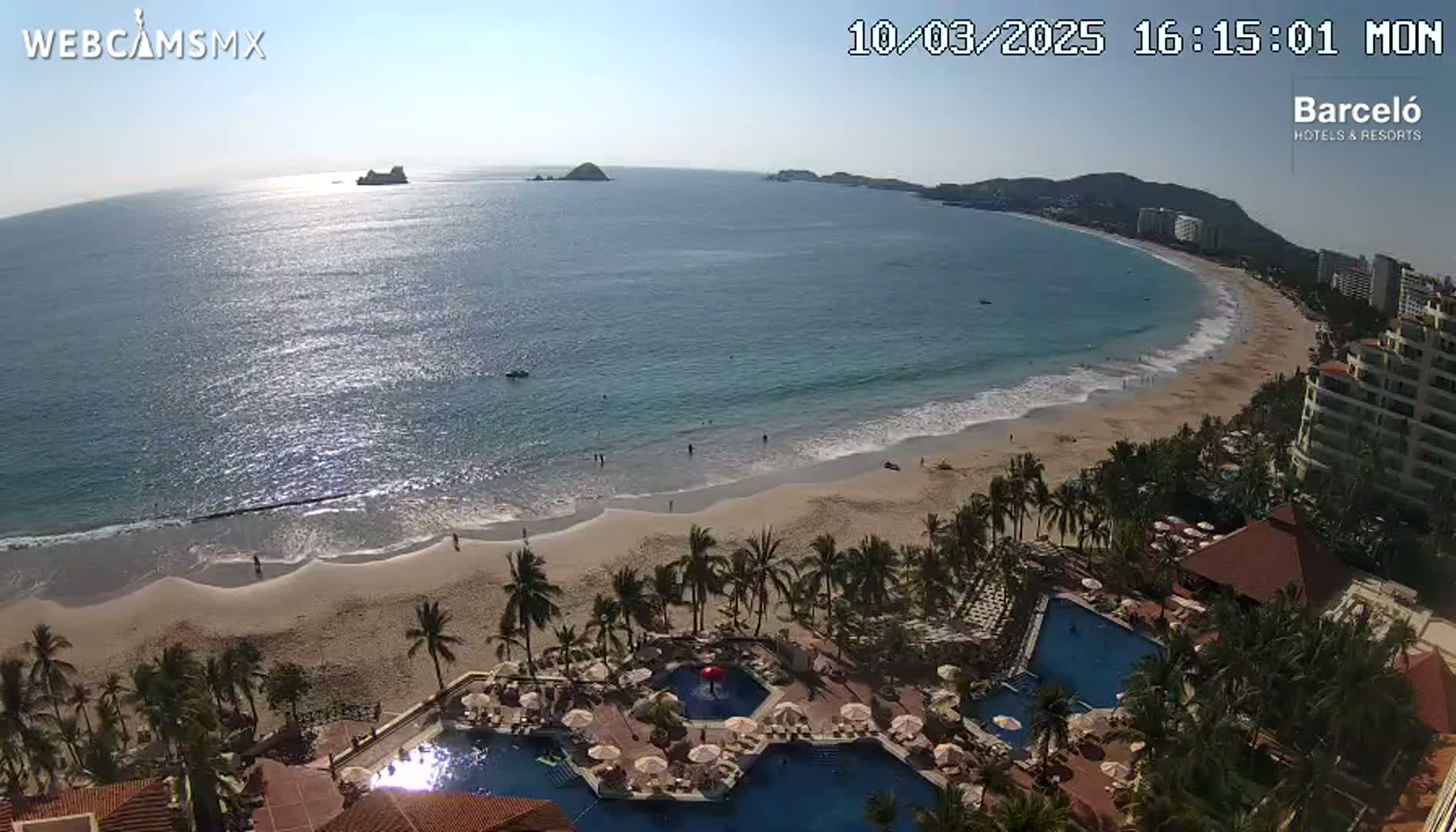 A sunny beach with calm waves, palm trees, and a resort with multiple pools is shown from an elevated viewpoint.