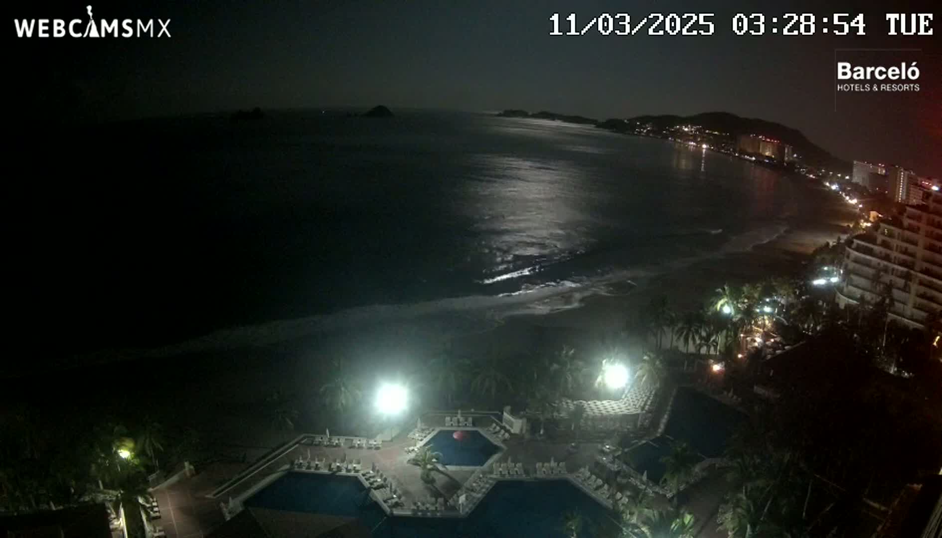 A nighttime view of a resort with multiple pools overlooking a dark ocean beach under a moonlit sky.
