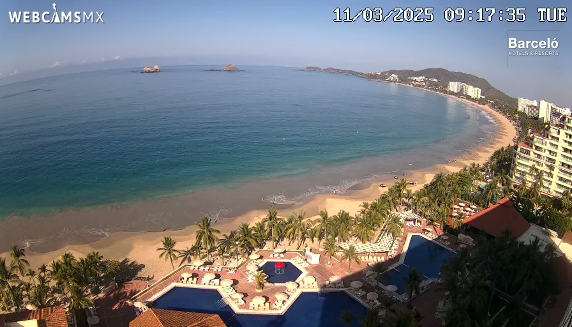 A beach resort with multiple pools is situated on a sandy beach next to a calm, turquoise ocean under a clear, sunny sky.