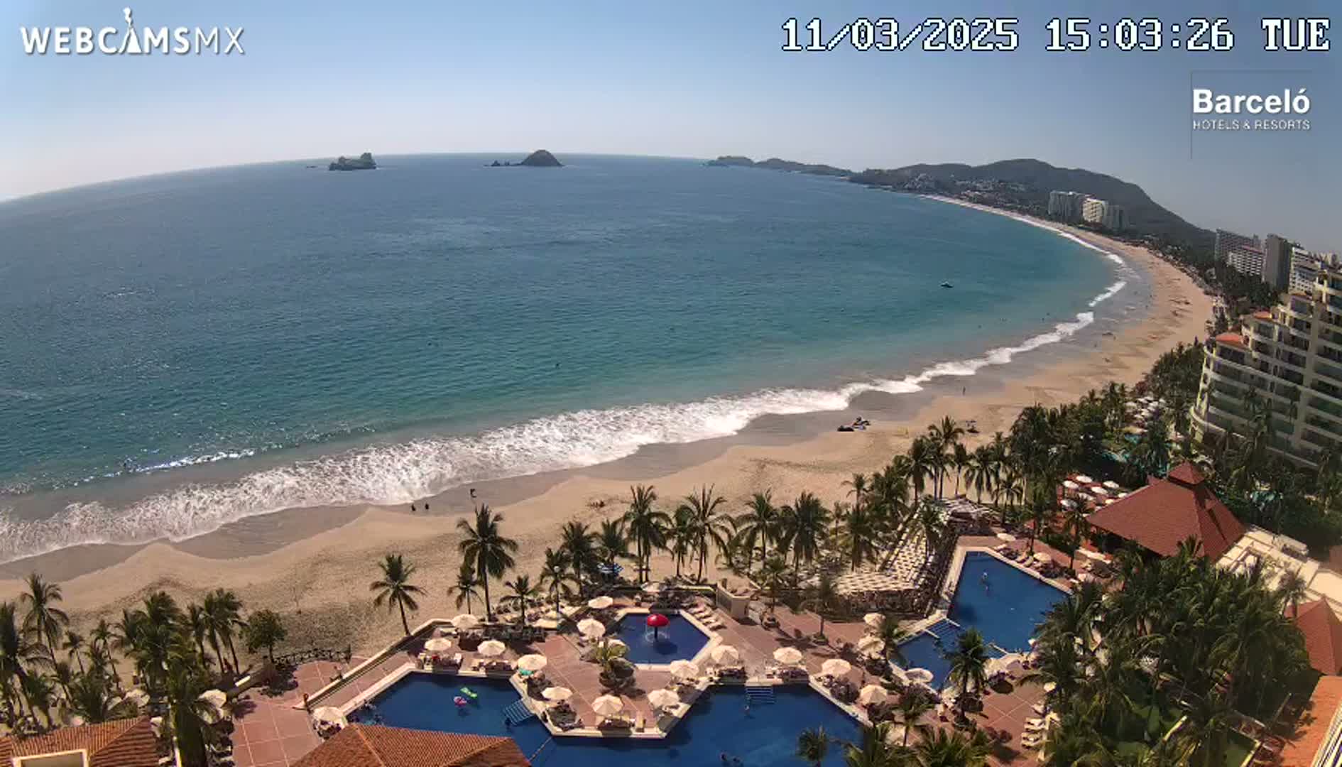 A beach with gentle waves laps a sandy shore, lined with palm trees and resort buildings under a clear blue sky.