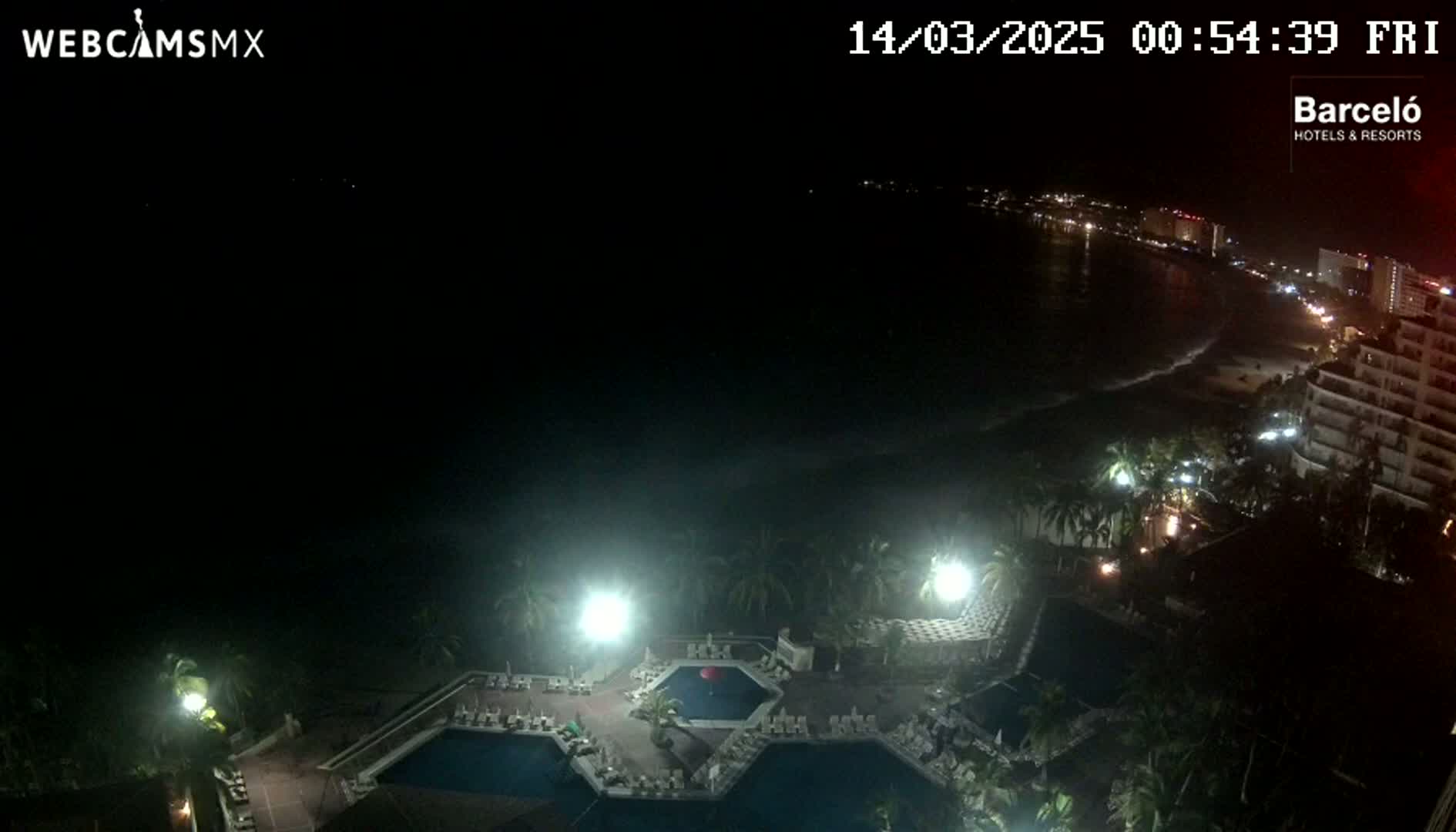 A nighttime, high-angle view shows a resort complex with illuminated pools and surrounding vegetation, overlooking a dark ocean and a dimly lit beach with buildings beyond.