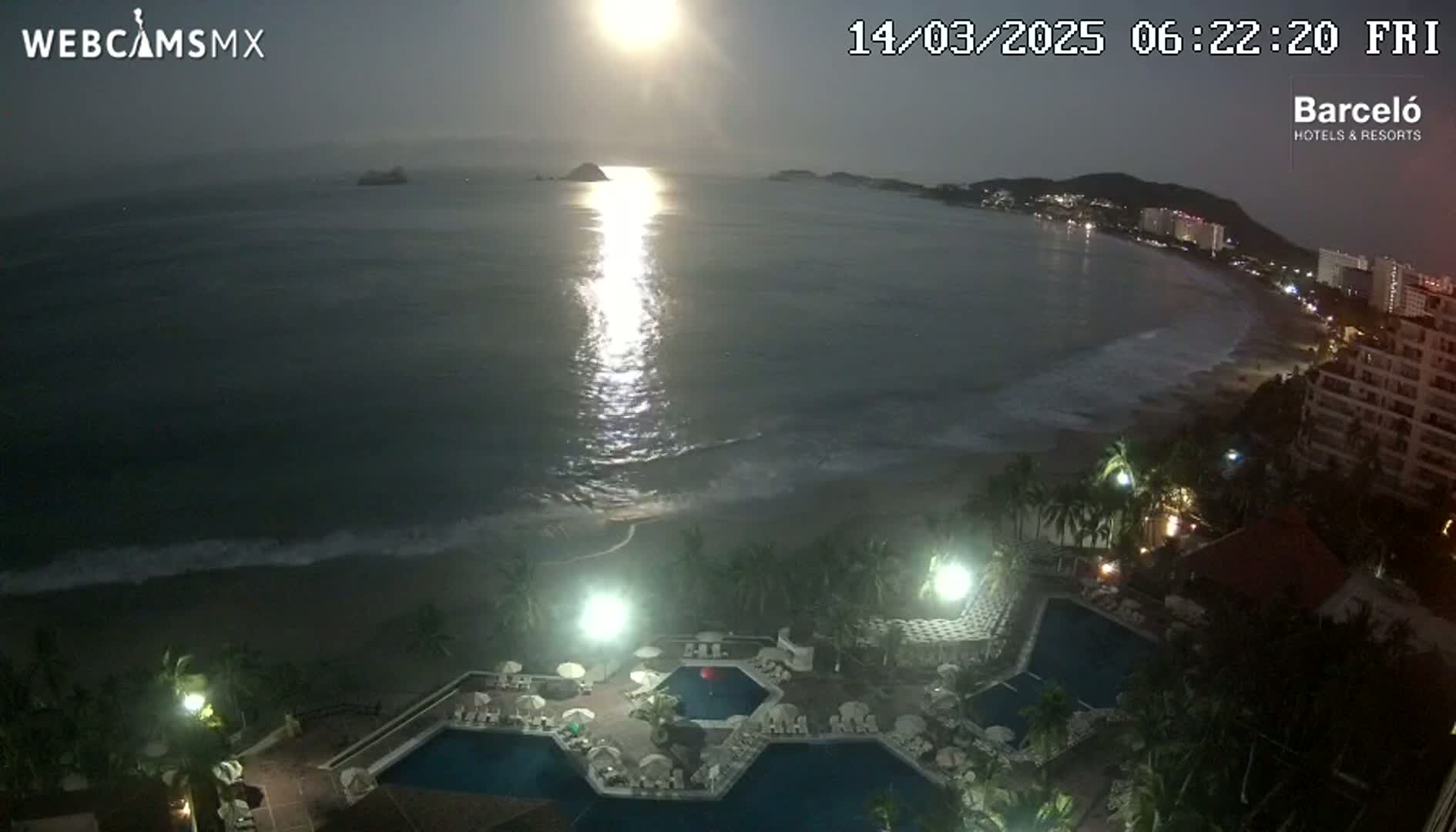 A nighttime view of a resort with multiple pools overlooking a beach and ocean under a bright moon.