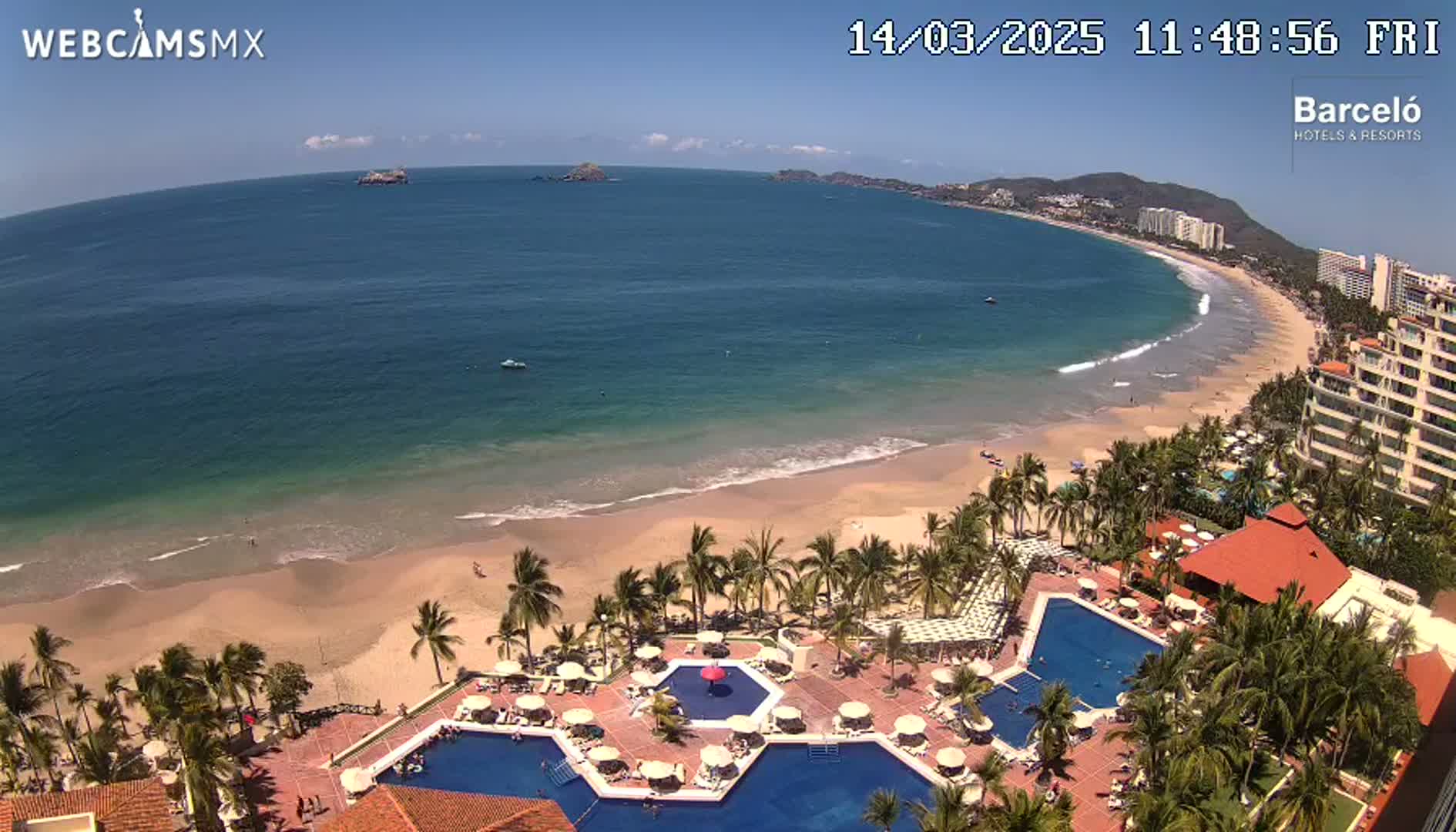An aerial view shows a resort with multiple pools, surrounded by palm trees and overlooking a sandy beach and a calm ocean on a sunny day.