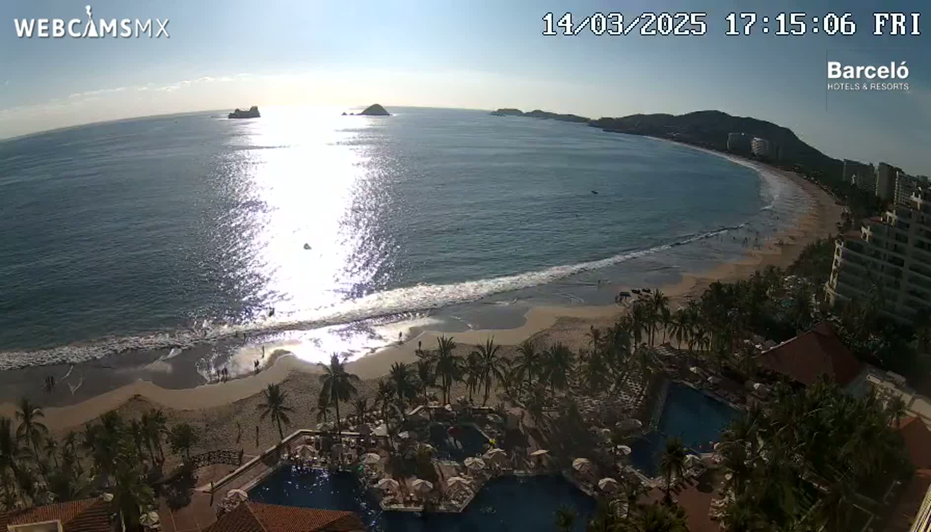 A sunny, high-angle view shows a long, sandy beach with gentle waves lapping the shore, palm trees lining the beach, and a resort with pools in the foreground.