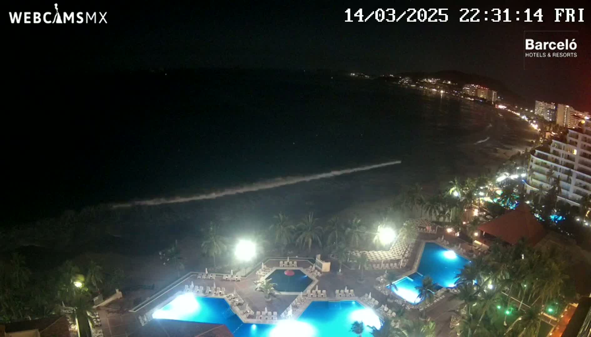 A nighttime view of a resort with illuminated pools, palm trees, and a beach with gently lapping waves under a dark sky.