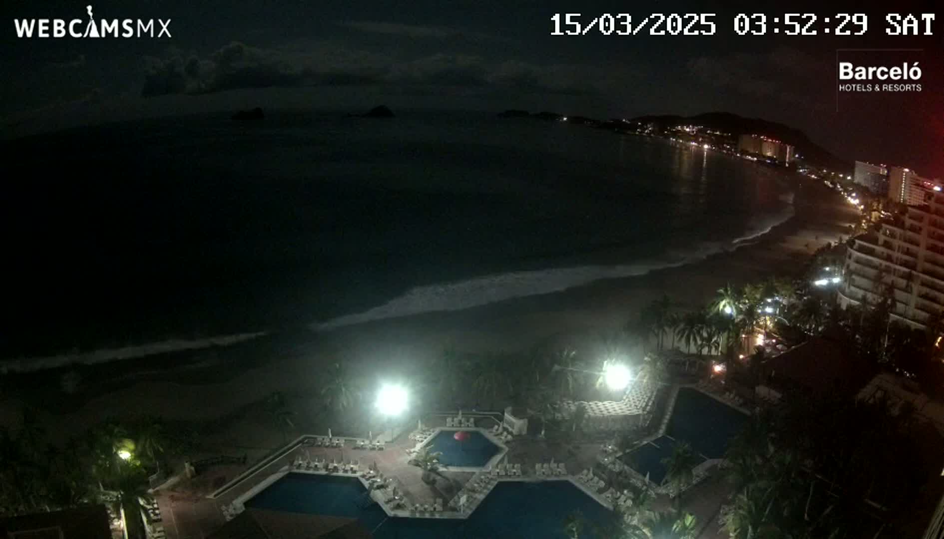 A nighttime coastal view shows a resort with several illuminated pools overlooking a dark ocean with gentle waves under a partly cloudy sky.