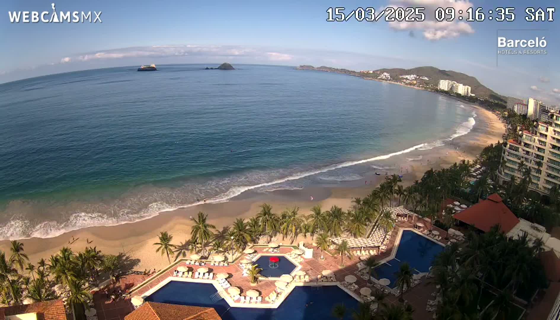 An aerial view shows a resort with multiple pools overlooking a sandy beach and ocean under sunny skies.