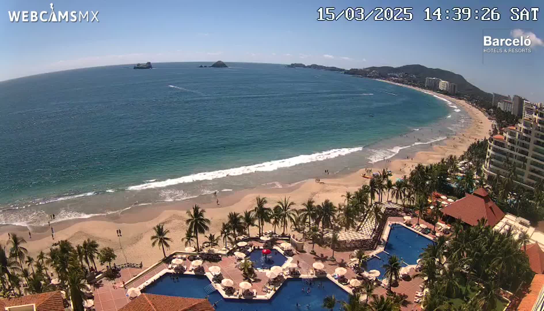An aerial view shows a beach with calm waves lapping a sandy shore, palm trees, a resort with multiple pools, and distant islands under a clear, sunny sky.