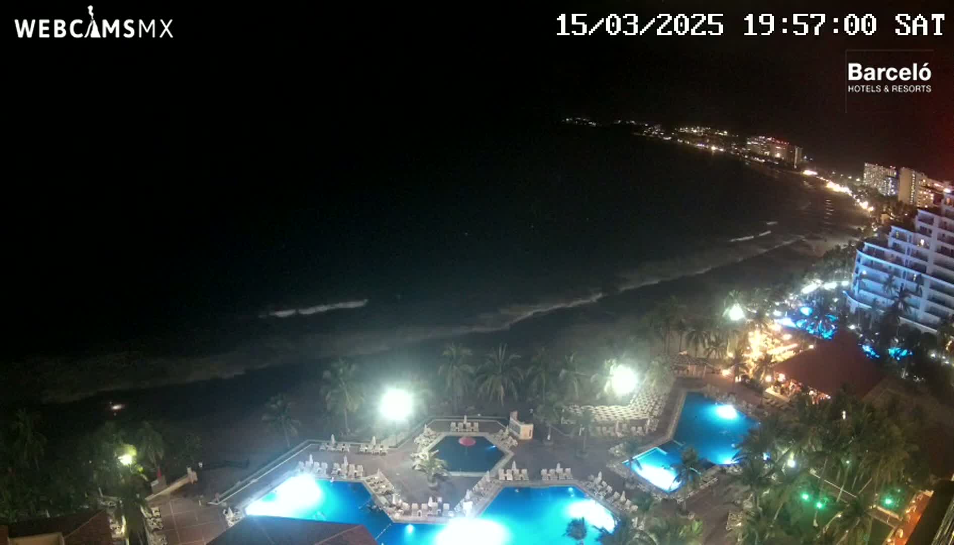 A nighttime view of a resort with multiple illuminated pools, situated on a beach with gently lapping waves under a clear, dark sky.