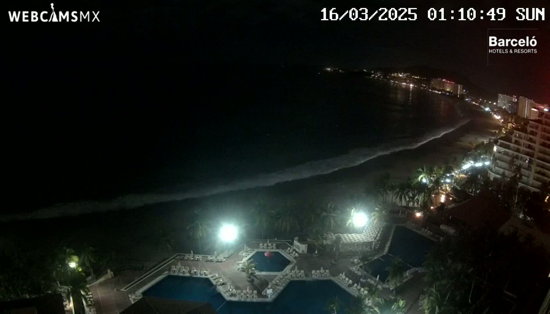 A nighttime view of a resort's pool area overlooking a dark ocean with waves lapping the shore and a dimly lit beach and cityscape in the distance.